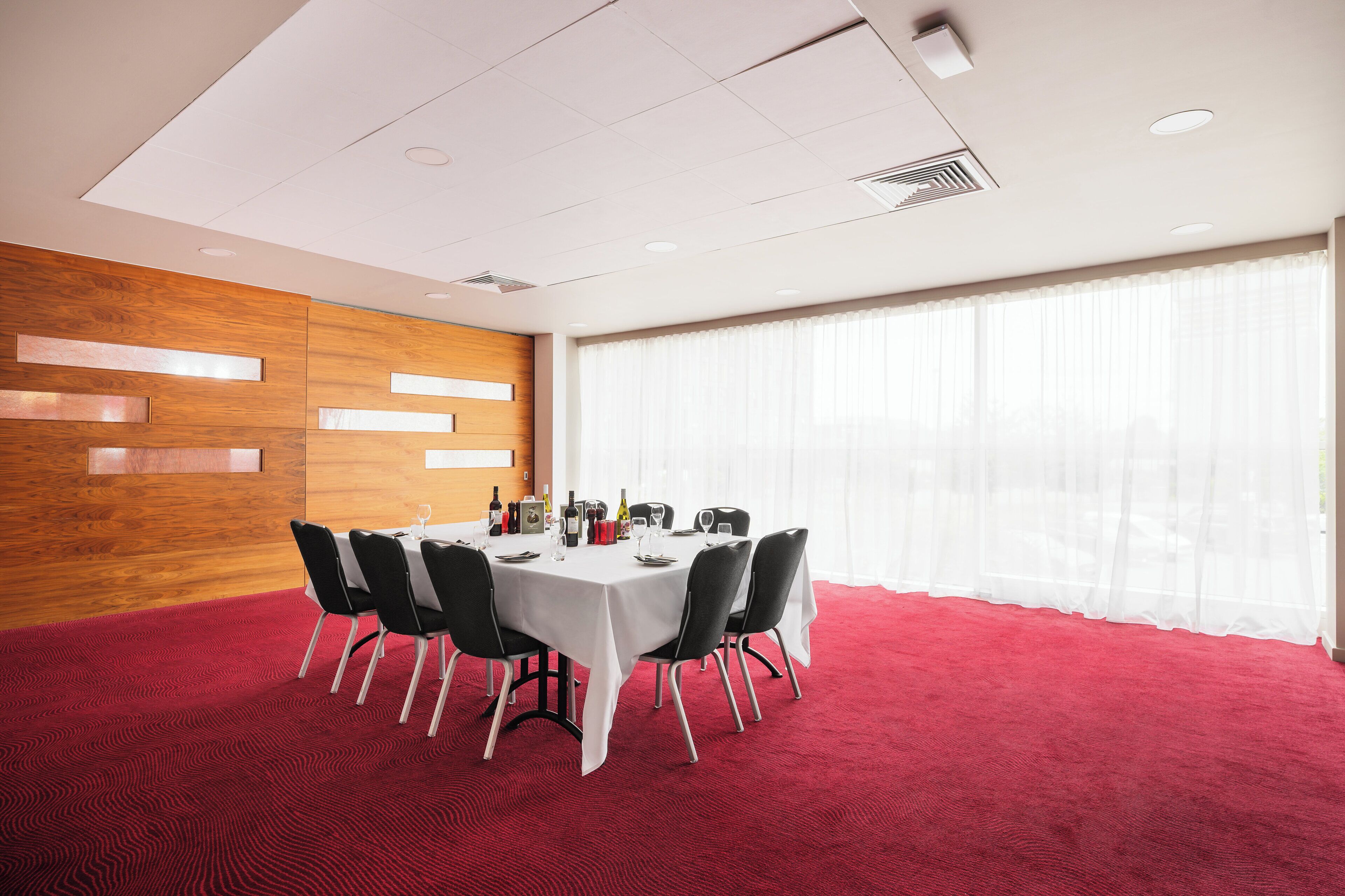 A conference room with a round table, black chairs, a red carpet, wooden panel walls, and large windows with sheer curtains.