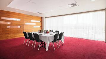 A conference room with a round table, black chairs, a red carpet, wooden panel walls, and large windows with sheer curtains.