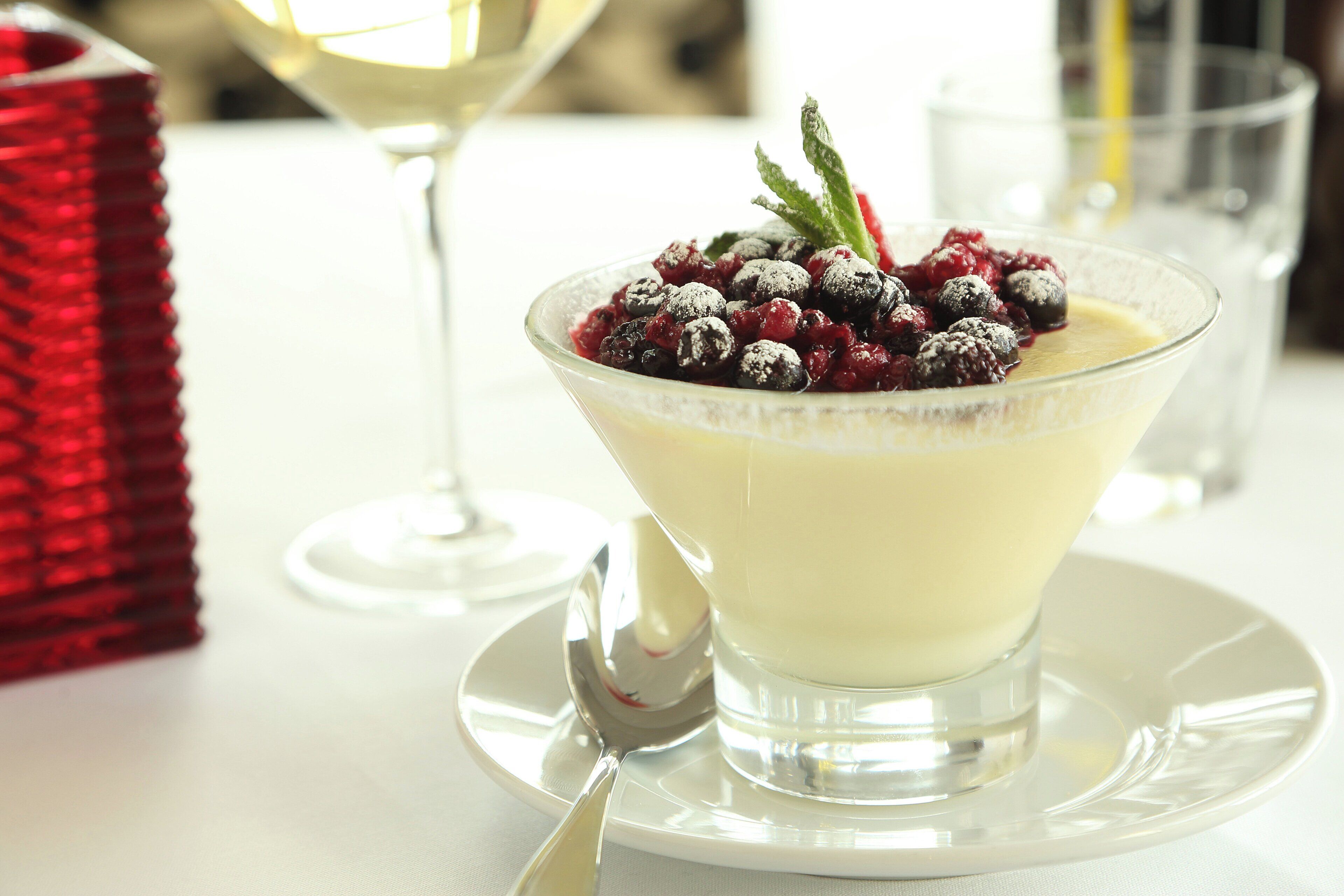 Dessert in einer klaren Glaskugel mit einem Löffel, Beeren und einer Minzdeko auf einem weißen Tisch.