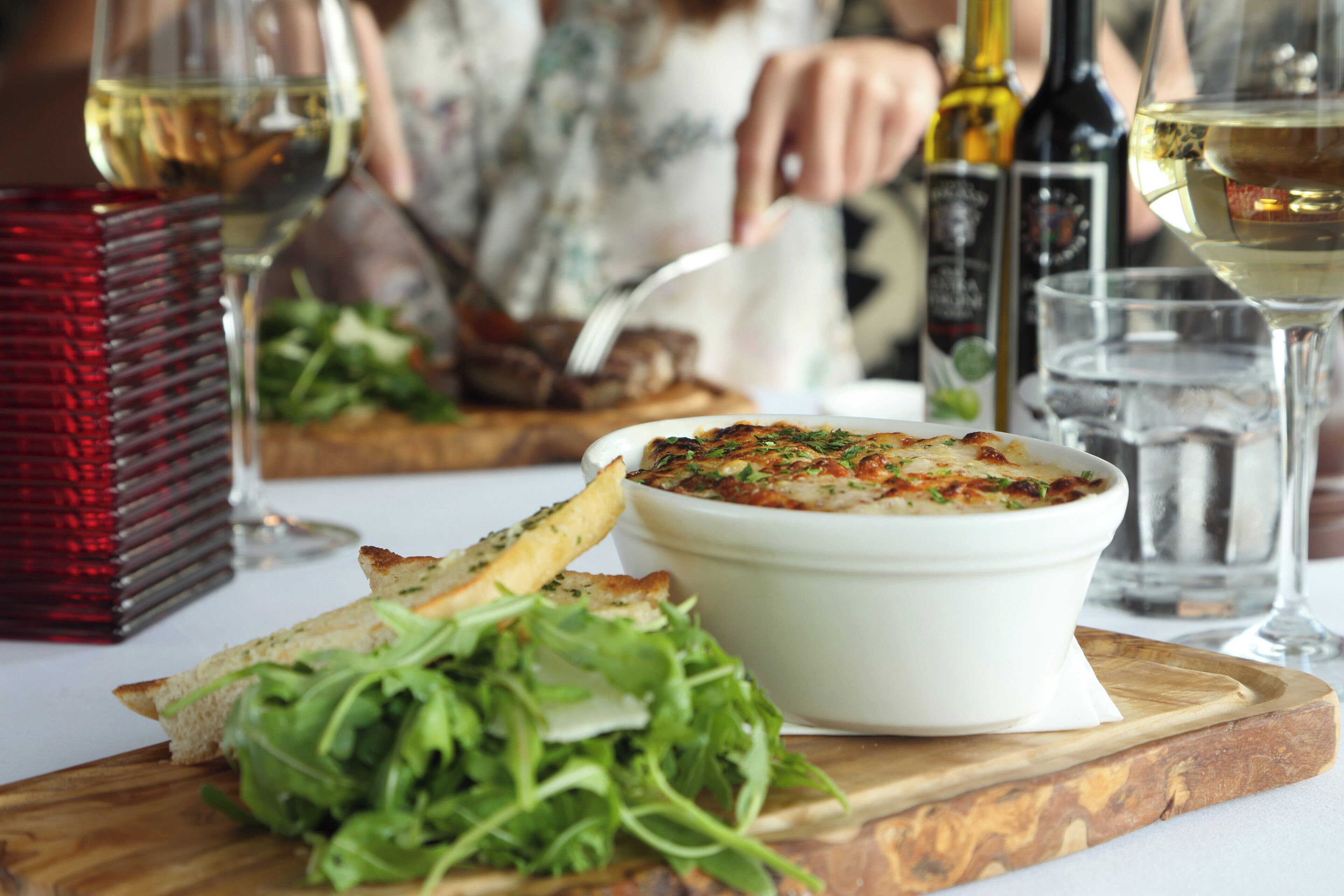 Une table à manger avec un bol blanc rempli de nourriture, une planche en bois avec du pain et des légumes verts, et des verres de vin blanc.