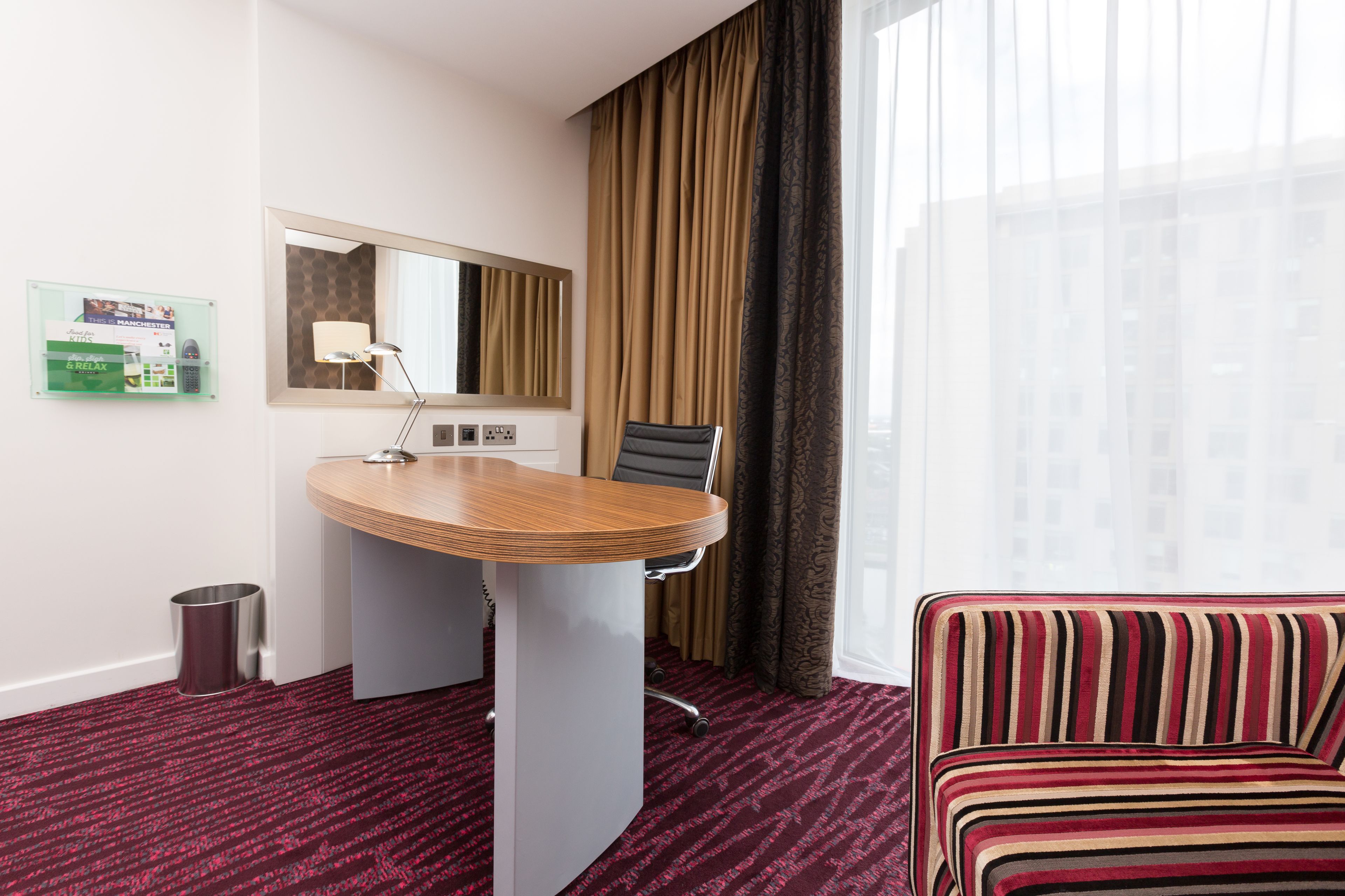 A hotel room with a wooden desk, a chair, a striped sofa, a large window with curtains, a mirror, and a bulletin board.