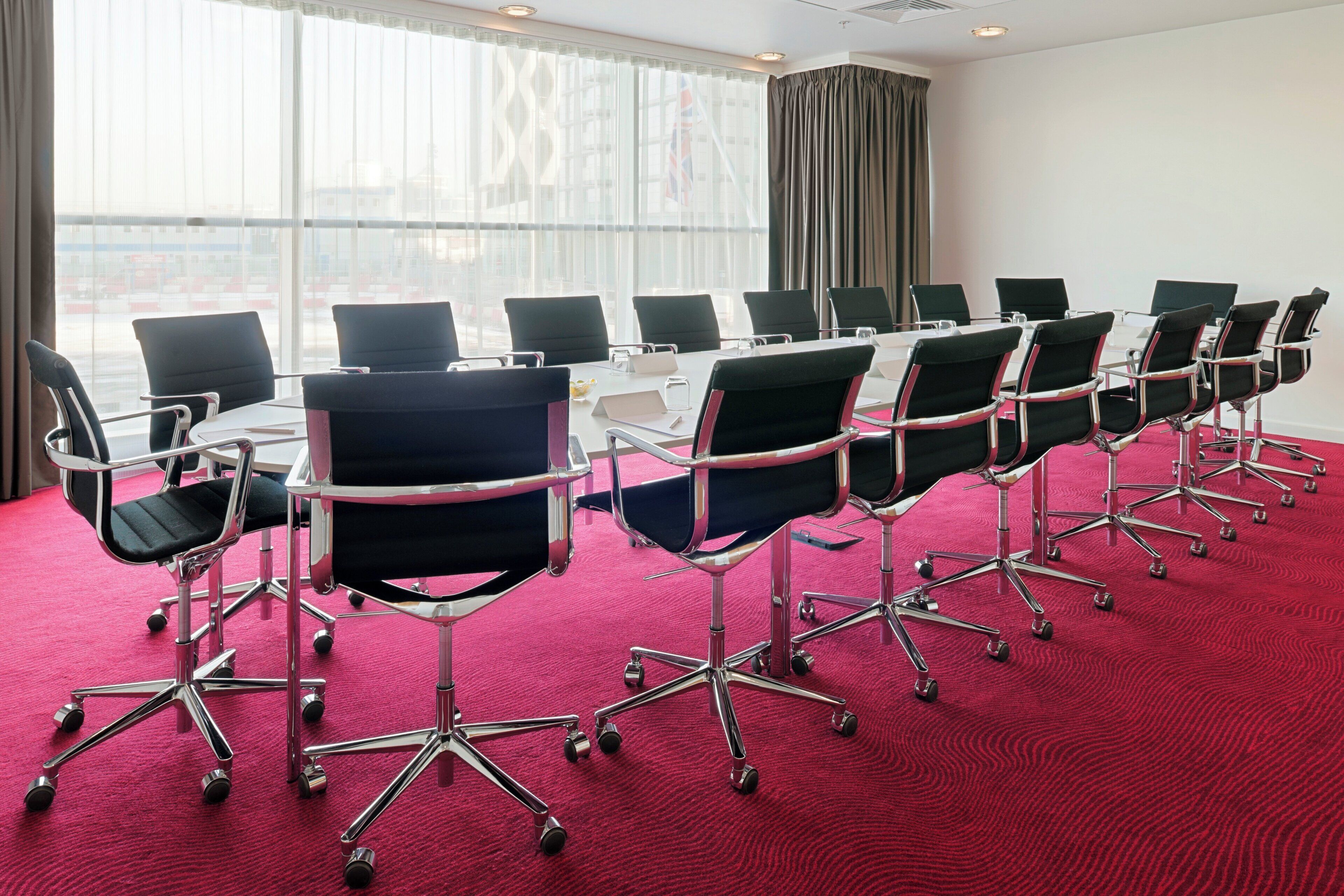 Une salle de conférence dotée d’une longue table et de nombreuses chaises, de grandes fenêtres avec des rideaux et d’un sol recouvert d’une moquette rouge.