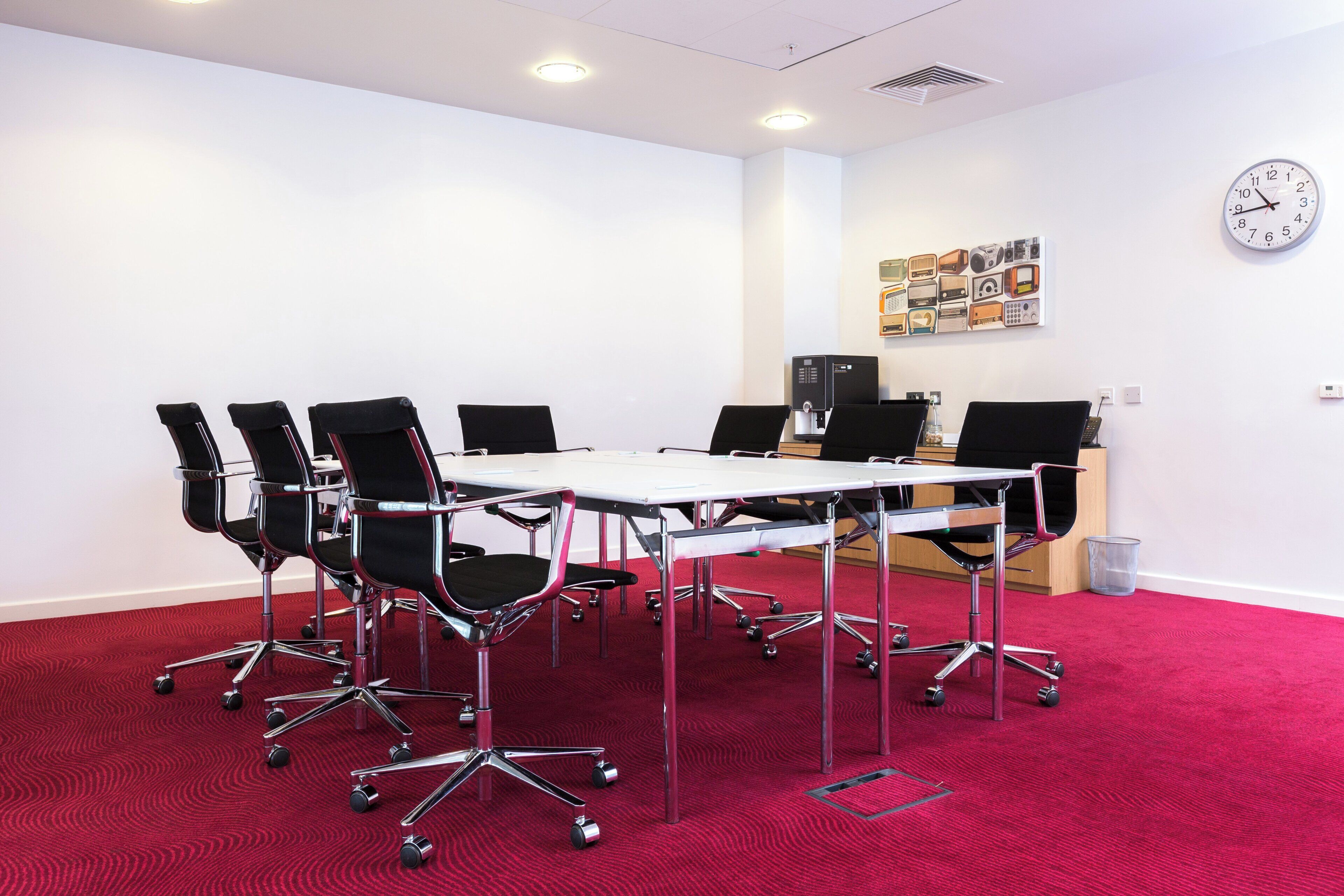 A conference room with a white table, black chairs, a clock, and a wall art piece.
