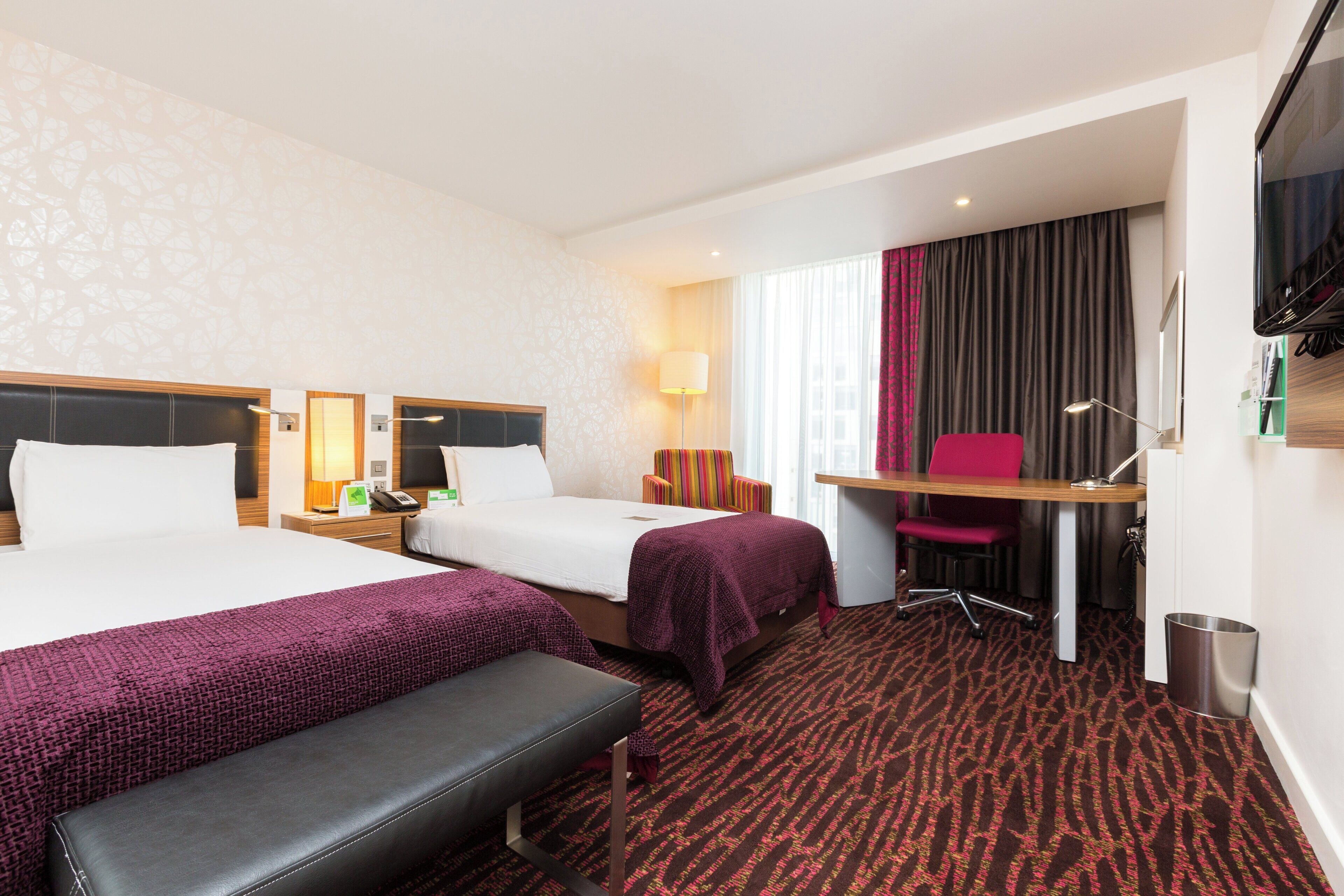 A hotel room with two beds, a desk, a chair, a bench, and a TV.