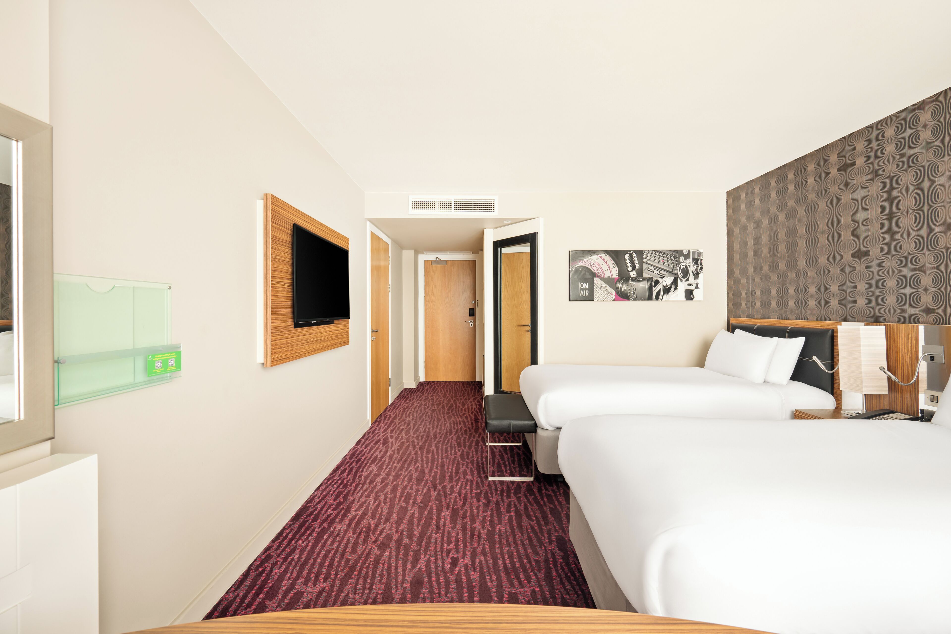 A hotel room with two beds, a television, a wall-mounted artwork, and a carpeted floor.