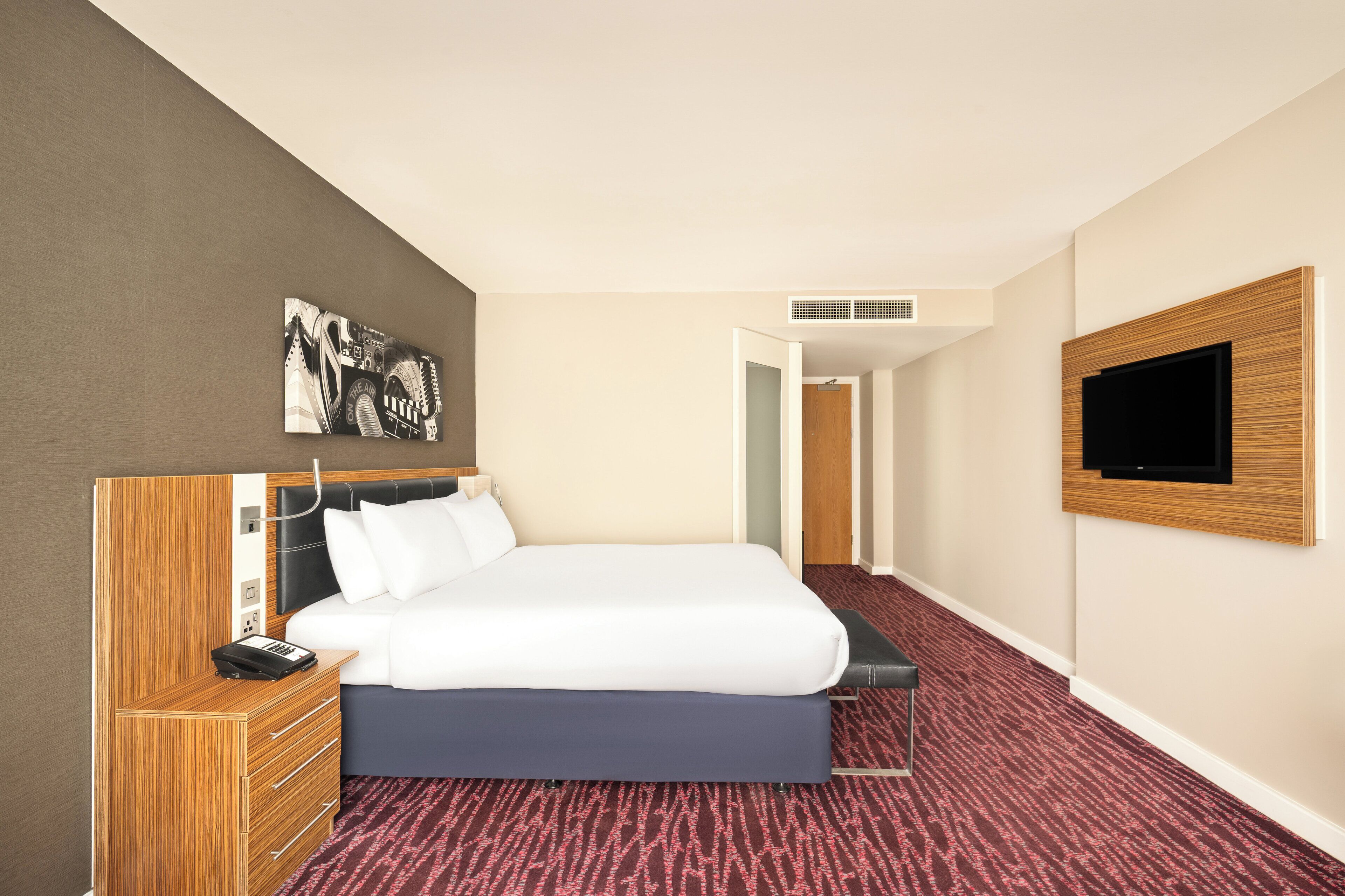 A hotel room with a bed, a desk with a telephone, a television mounted on the wall, and a door leading to another room.