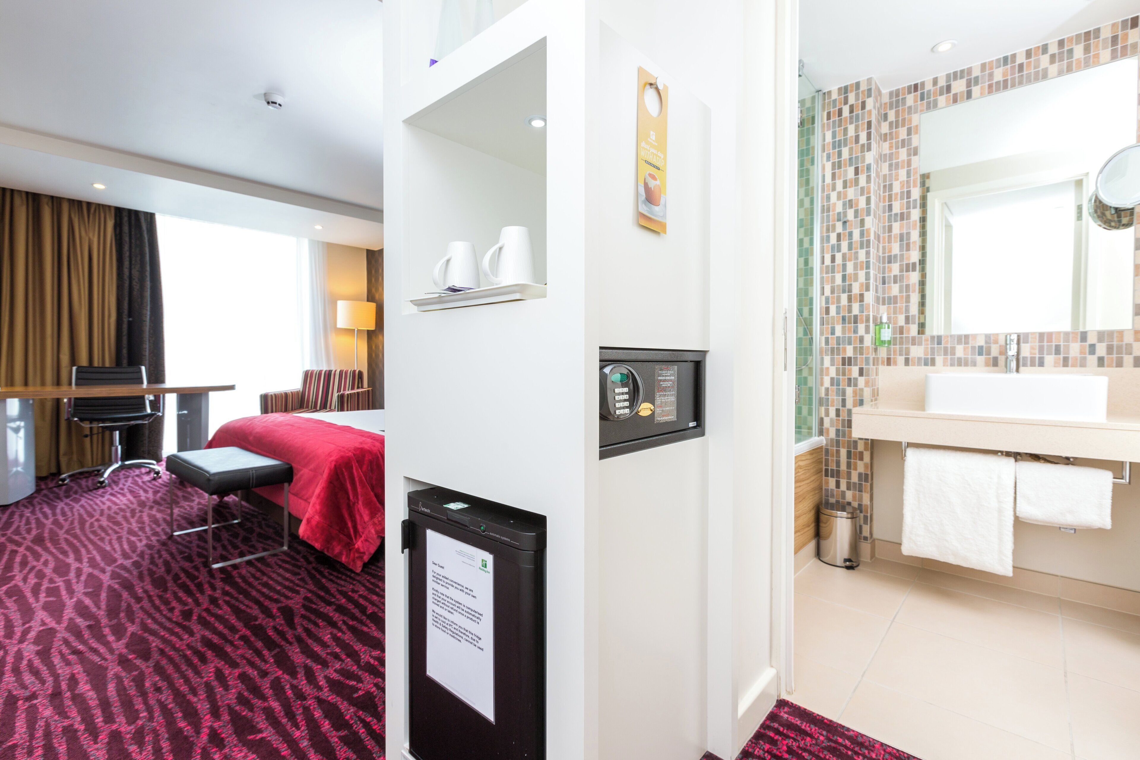 Une chambre d’hôtel comprenant un lit, un bureau, une chaise, une petite table, une poubelle et une salle de bain avec un lavabo et un miroir.