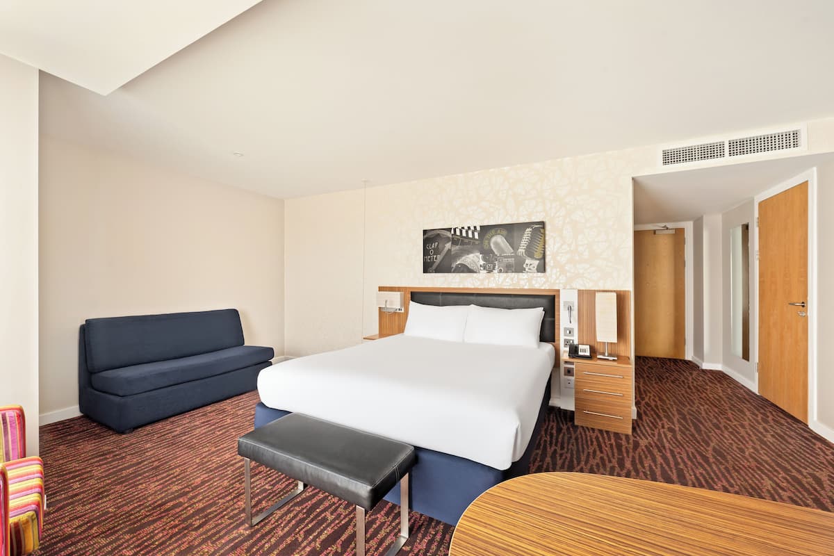 Una habitació d'hotel amb un llit, un sofà, una banca, un escriptori i una obra d'art penjada a la paret.