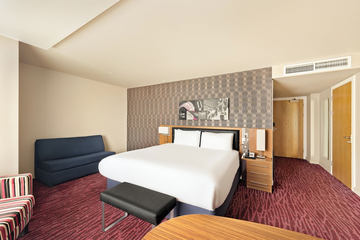 Habitación de hotel con cama, escritorio, silla, banco, mesita de noche y armario.