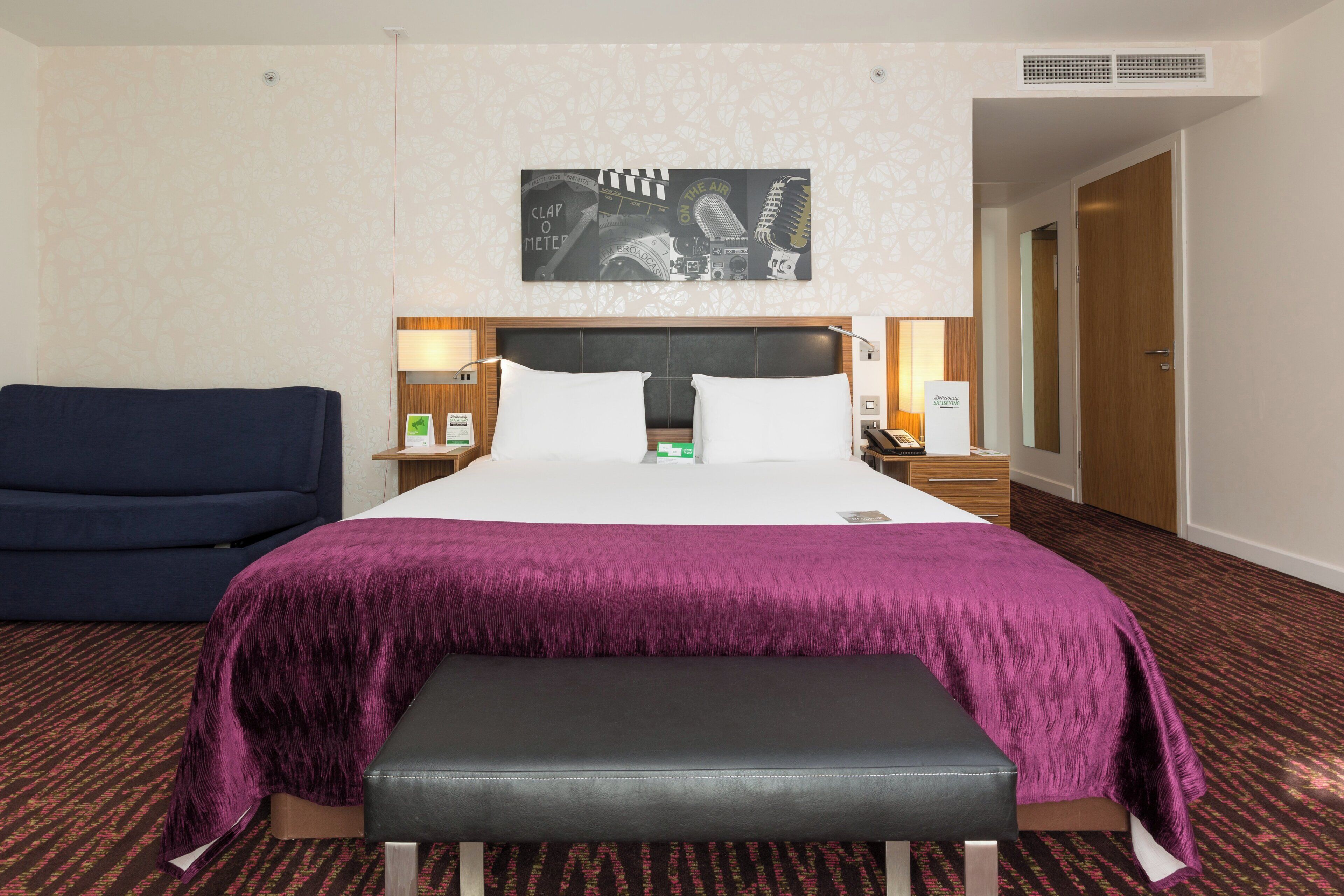 A hotel room with a bed, a bench, a nightstand, a lamp, and a wall-mounted artwork.