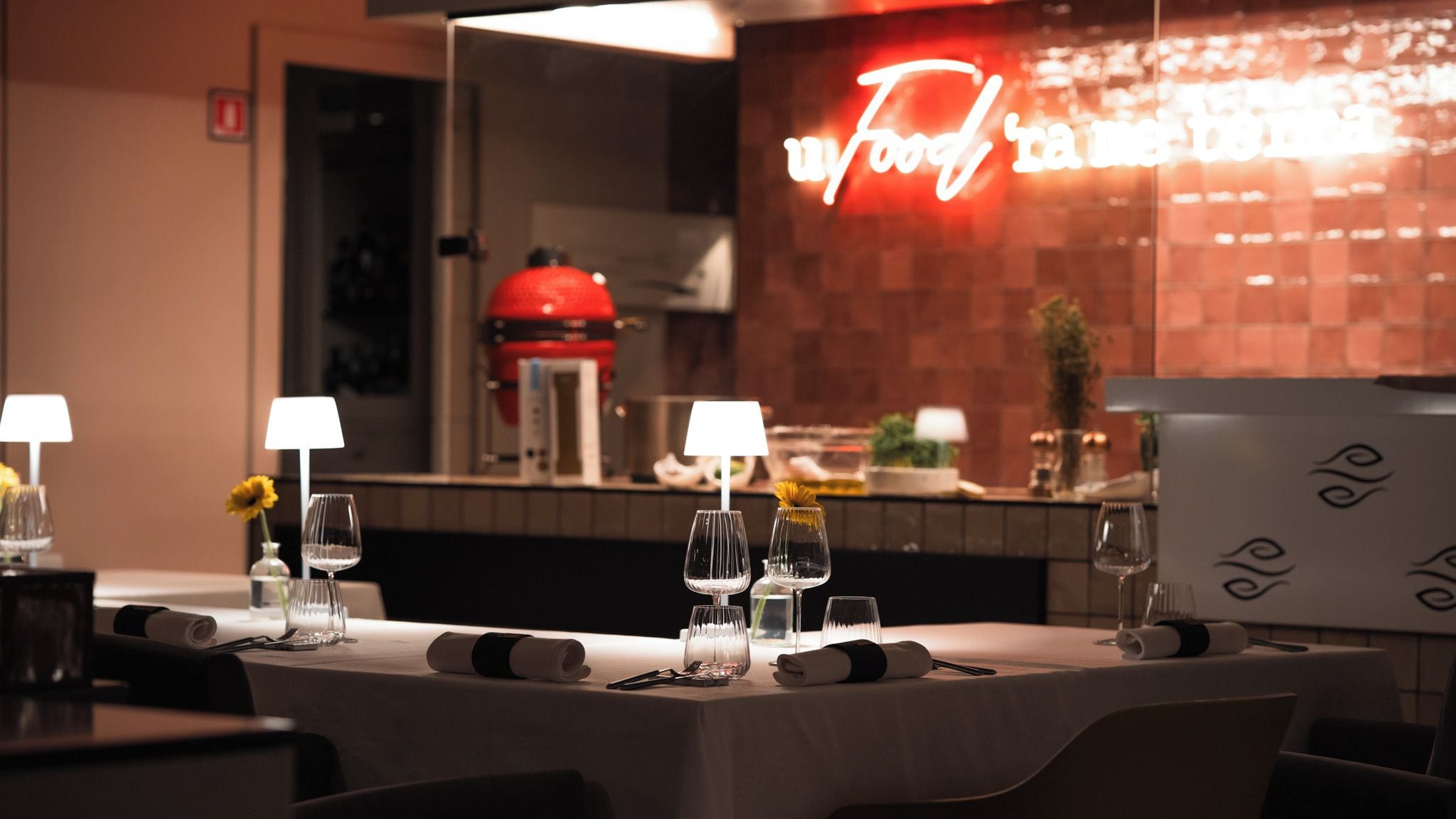 A restaurant interior with tables set for dining, a neon sign in the background, and a kitchen area visible through a glass partition.