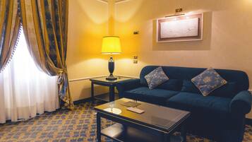 Une chambre d’hôtel avec un canapé bleu, une petite table, une lampe, une fenêtre avec des rideaux et un tableau accroché au mur.