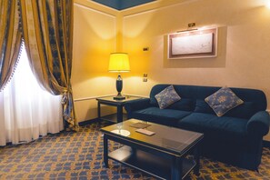 A hotel room with a blue sofa, a small table, a lamp, a window with curtains, and a wall-mounted picture. - Altafiumara Resort & Spa (Villa San Giovanni)