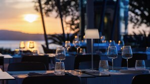 A restaurant setting with tables set for dining, a lamp, and a view of the sunset over the water. - Altafiumara Resort & Spa (Villa San Giovanni)