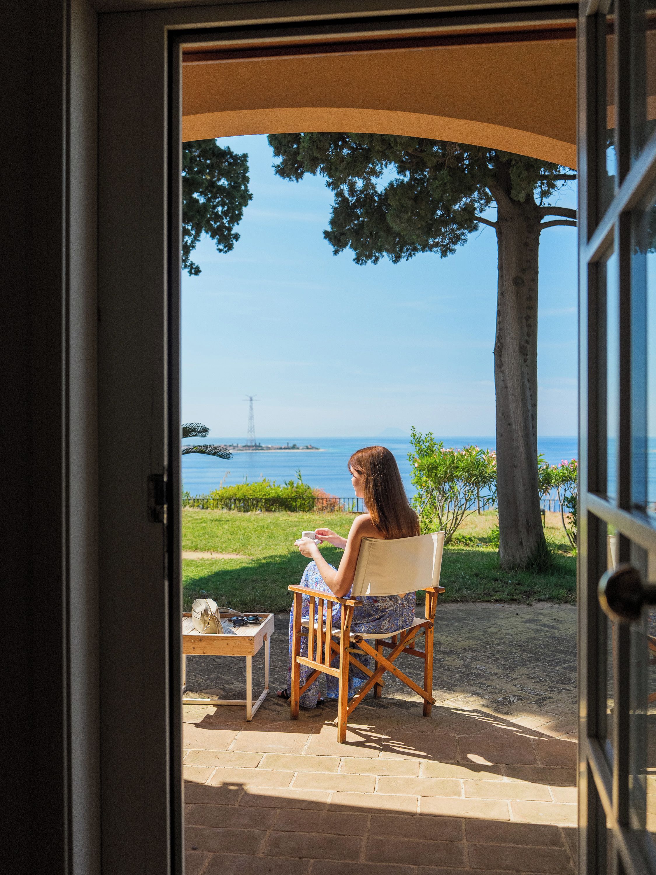A person seated in a chair with a view of the sea and a tree.