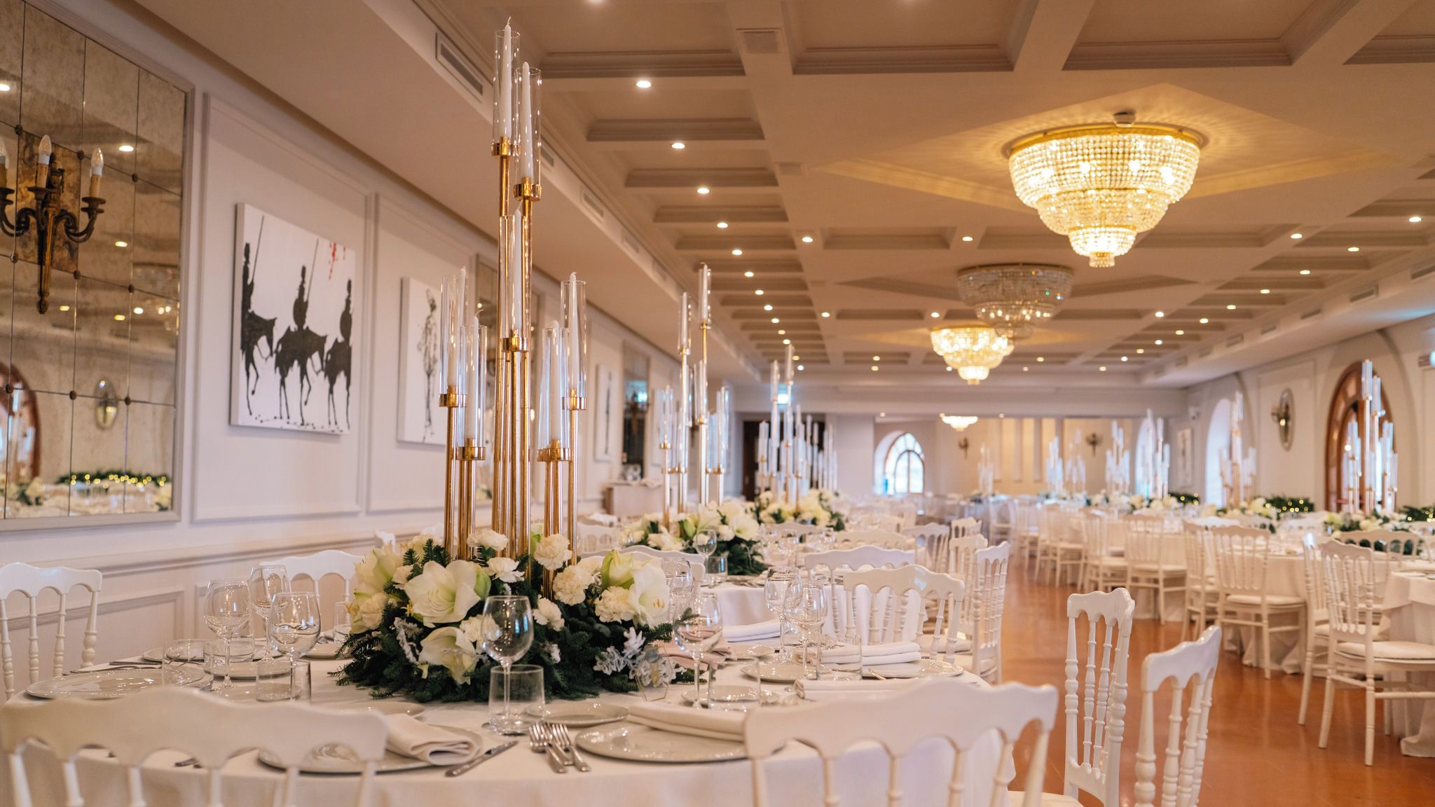 A banquet hall with long tables set for a formal event, featuring tall candle holders, white floral centerpieces, and elegant chandeliers.