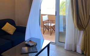 A room with a sofa, a coffee table with books, a chair, and a sliding glass door leading to an outdoor area with a chair. - Altafiumara Resort & Spa (Villa San Giovanni)