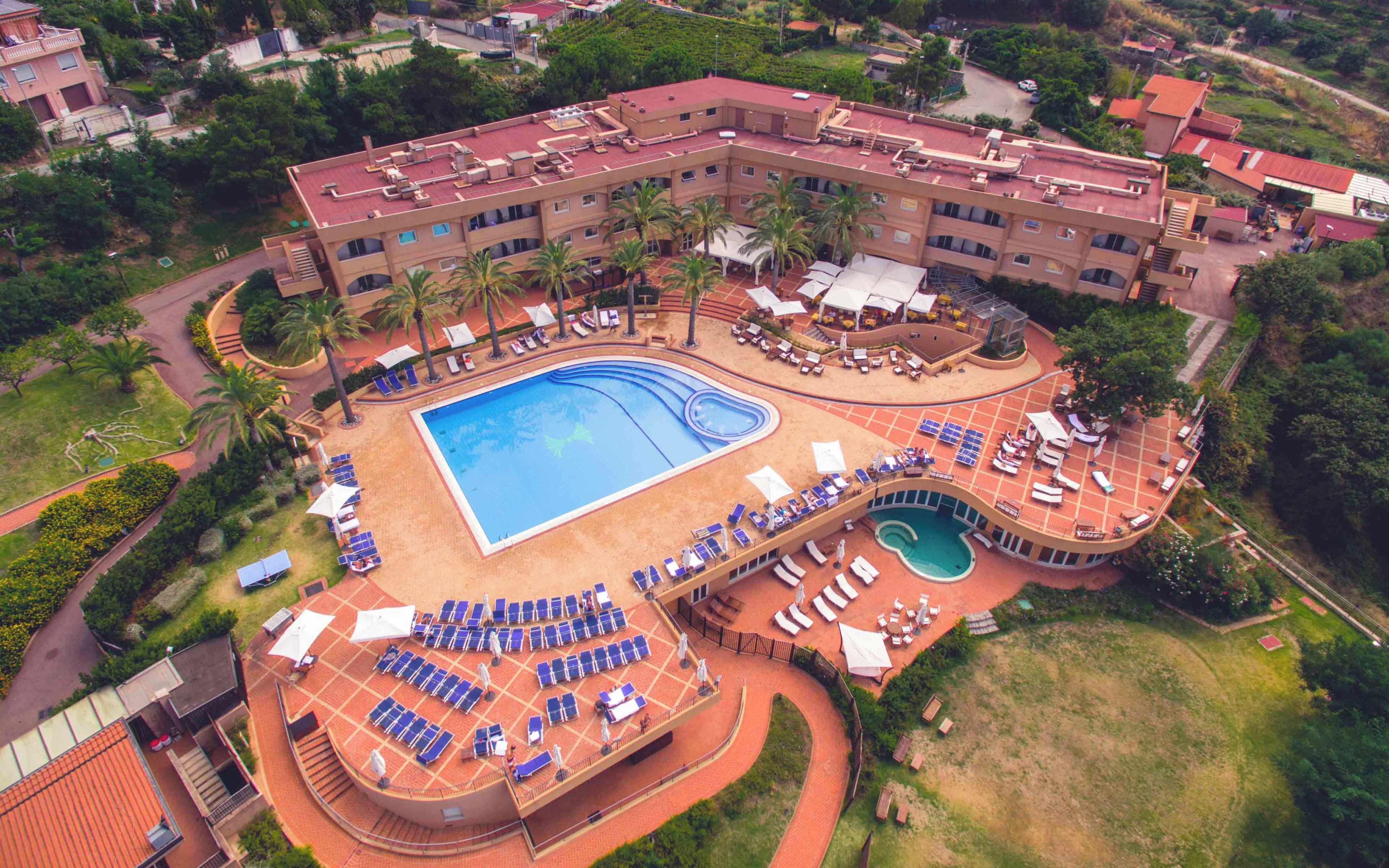 A large hotel with a central swimming pool, multiple sunbathing areas, and a surrounding landscaped garden.