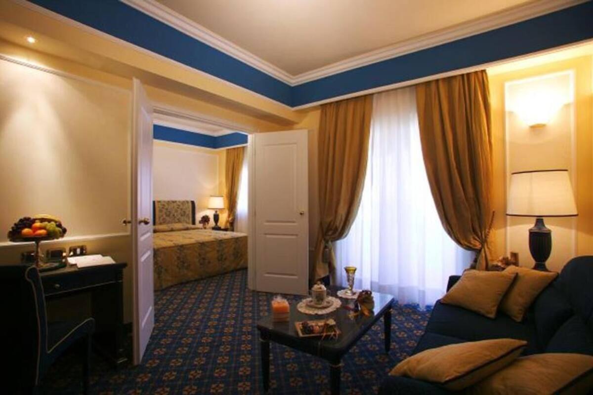 A hotel room with a bed, a desk with a chair, a sofa, and a dining table with a fruit bowl and a lamp.