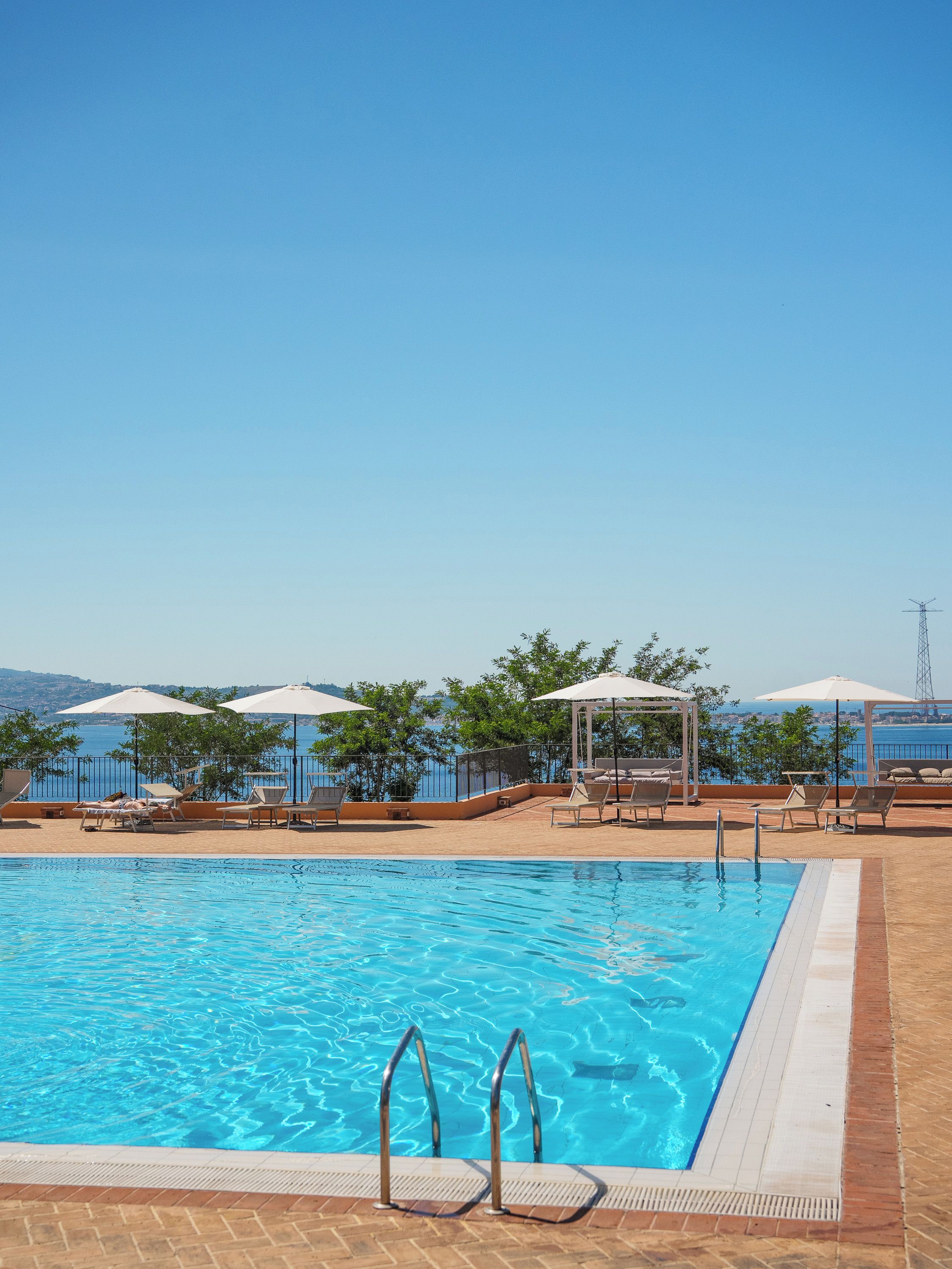 Piscina con sillas de descanso y sombrillas, rodeada de una valla y árboles, con vista a una masa de agua.