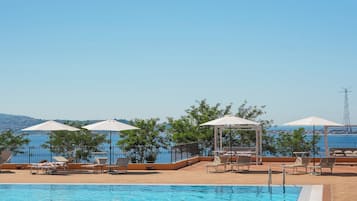 Piscina con sillas de descanso y sombrillas, rodeada de una valla y árboles, con vista a una masa de agua.
