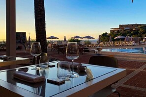 A dining area with a table set for two, overlooking a pool and beach umbrellas. - Altafiumara Resort & Spa (Villa San Giovanni)
