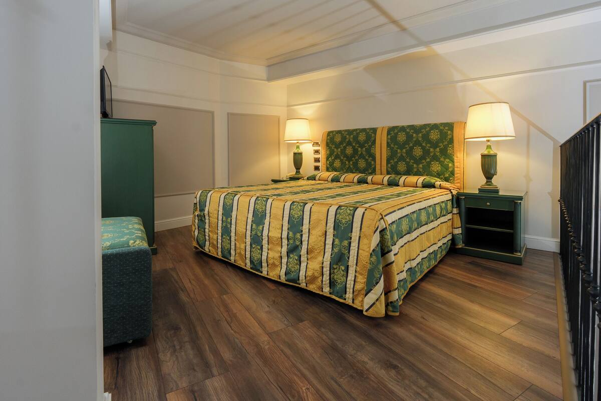 A bedroom with a large bed, two bedside tables with lamps, a green cabinet, and a wooden floor.
