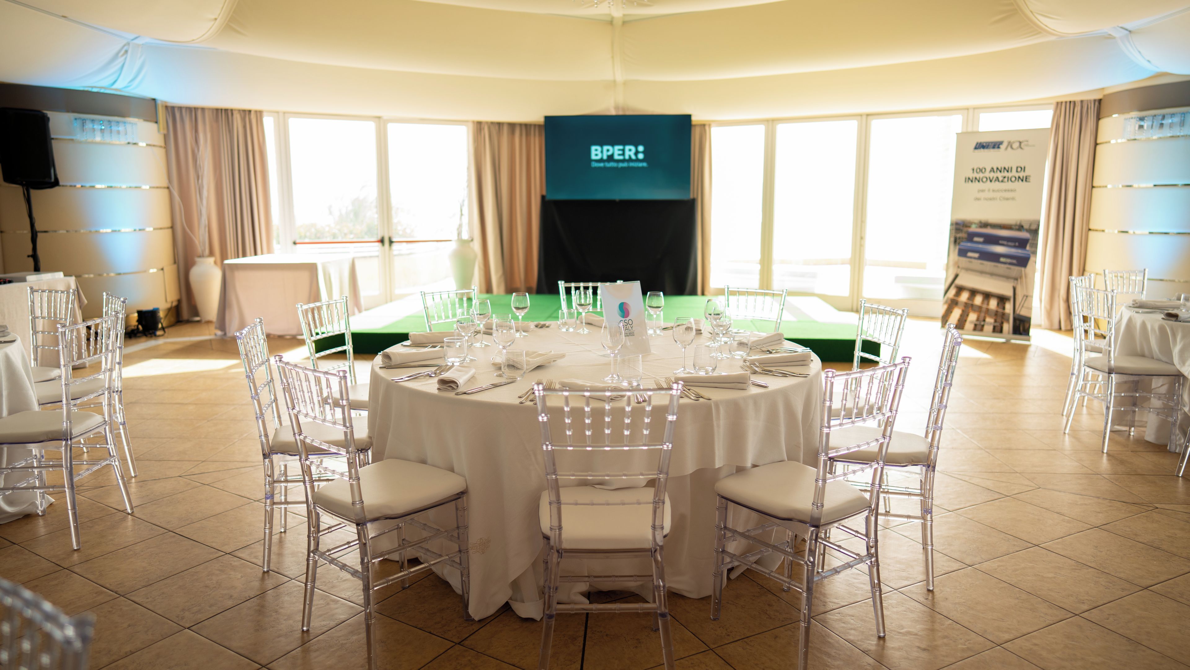 A round table set for a formal event with clear chairs, a white tablecloth, and a banner in the background.