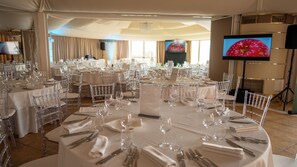 A banquet hall with round tables set for a formal event, clear chairs, and a large screen displaying an image of an apple. - Altafiumara Resort & Spa (Villa San Giovanni)
