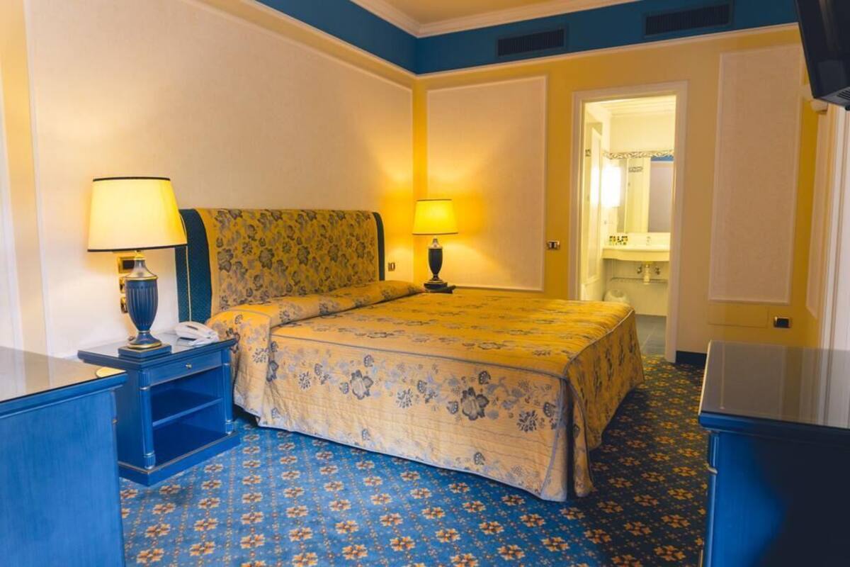 A hotel room with a bed, two bedside tables with lamps, a blue carpet, and a bathroom visible through an open door.