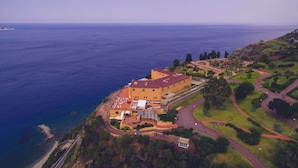 Vue aérienne d’un hôtel situé sur la côte, avec un grand bâtiment, une plage et un jardin aménagé.