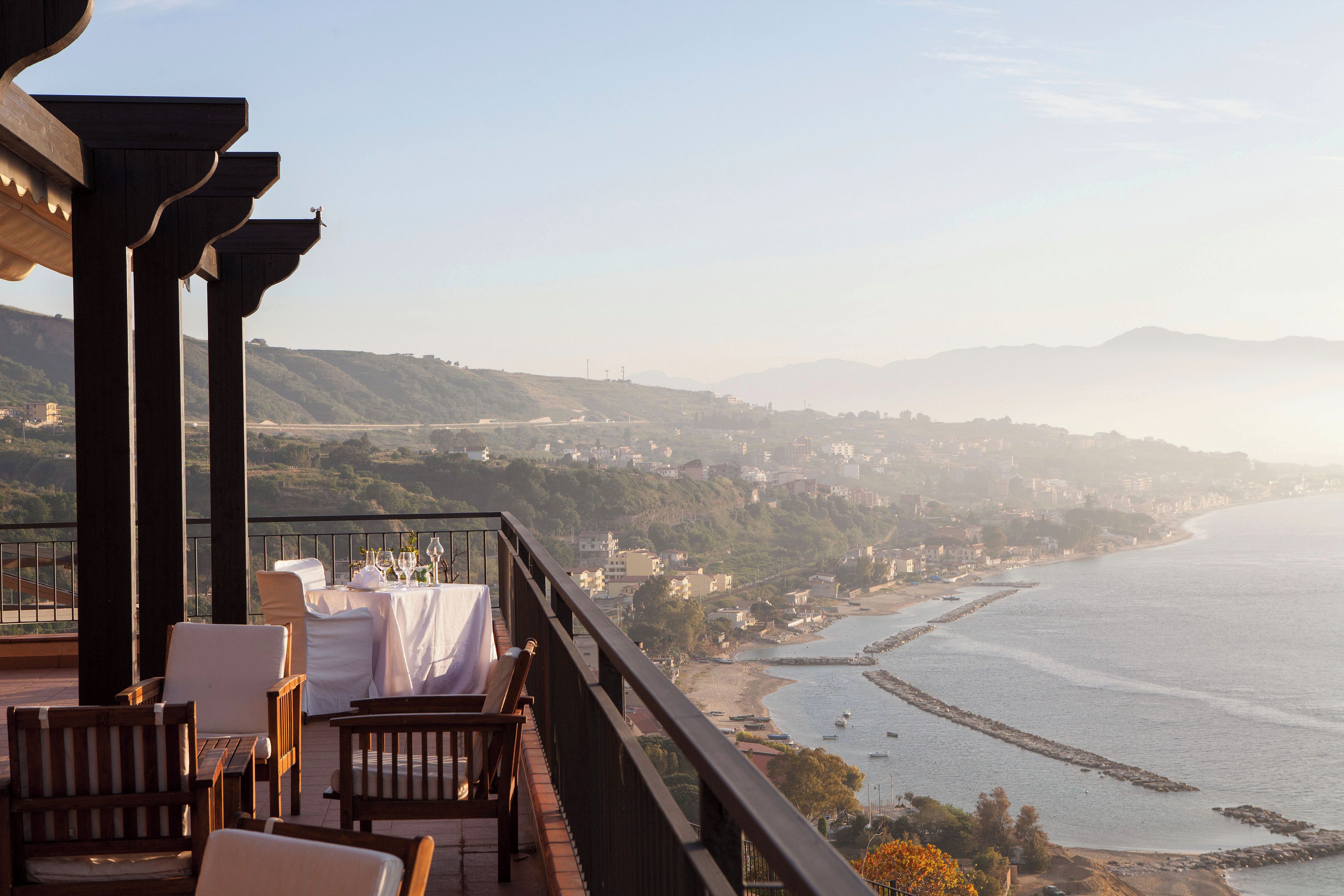A terrace with wooden furniture and a table set for dining, overlooking a coastal landscape with hills and a beach.