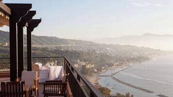 A terrace with wooden furniture and a table set for dining, overlooking a coastal landscape with hills and a beach.