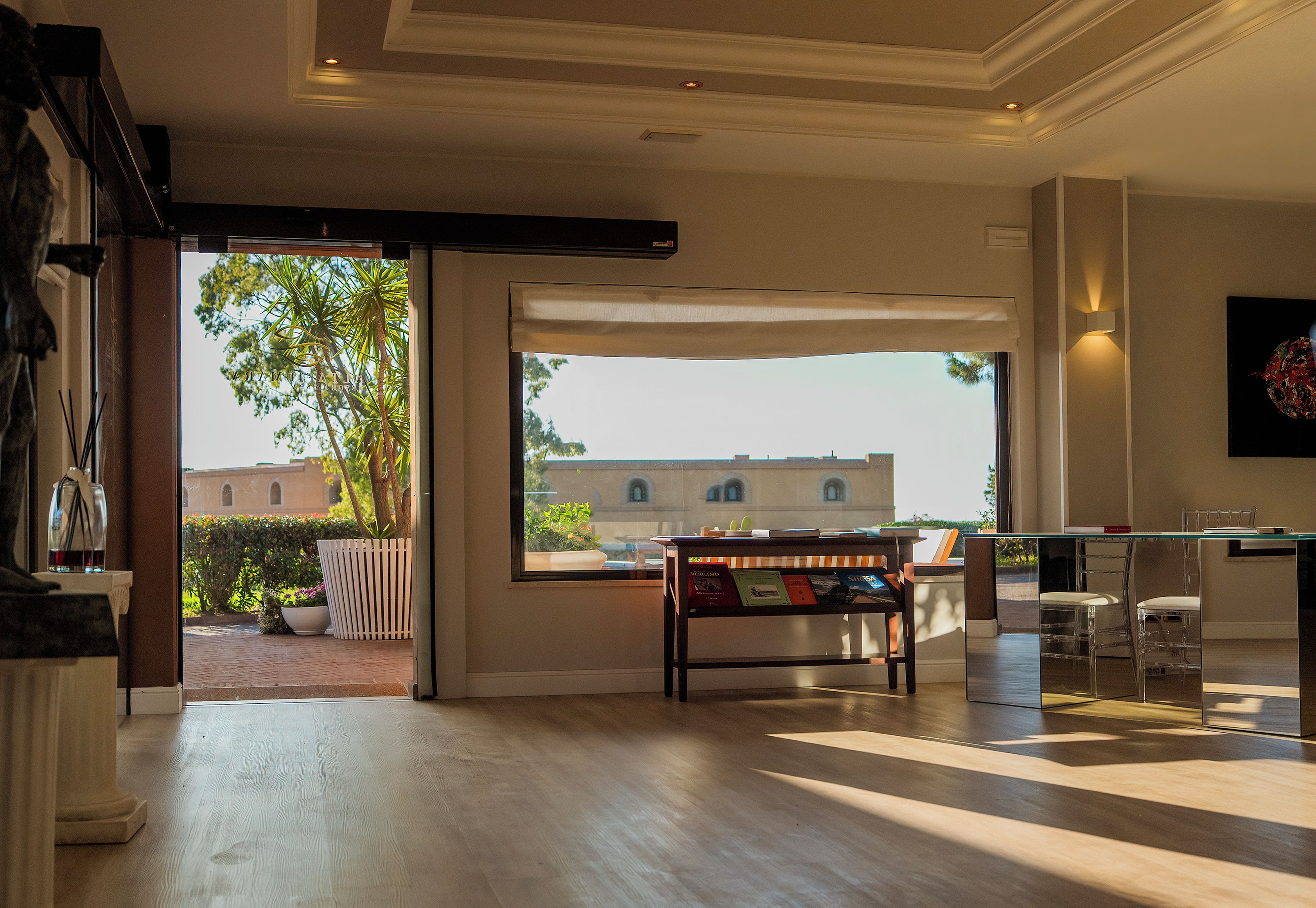 An open-plan living area with a view of a garden and building, a glass-top table with chairs, and a bookshelf.