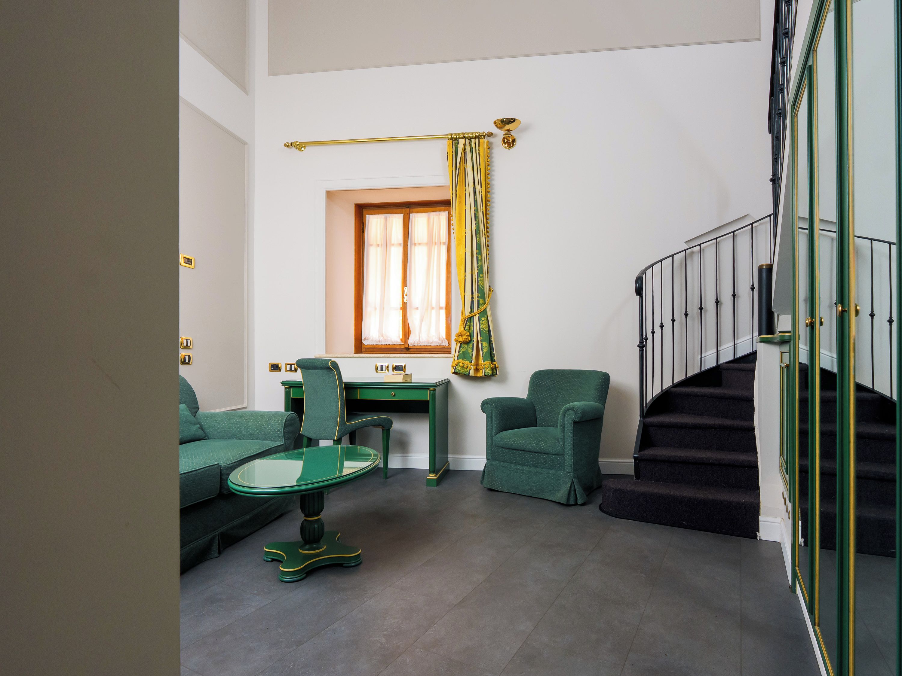 Una habitación con un sofá verde, una mesa de vidrio, un escritorio, una silla, una ventana con cortinas y una escalera con barandilla.