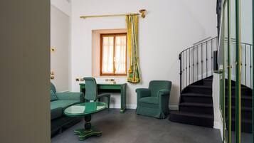 Una habitación con un sofá verde, una mesa de vidrio, un escritorio, una silla, una ventana con cortinas y una escalera con barandilla.