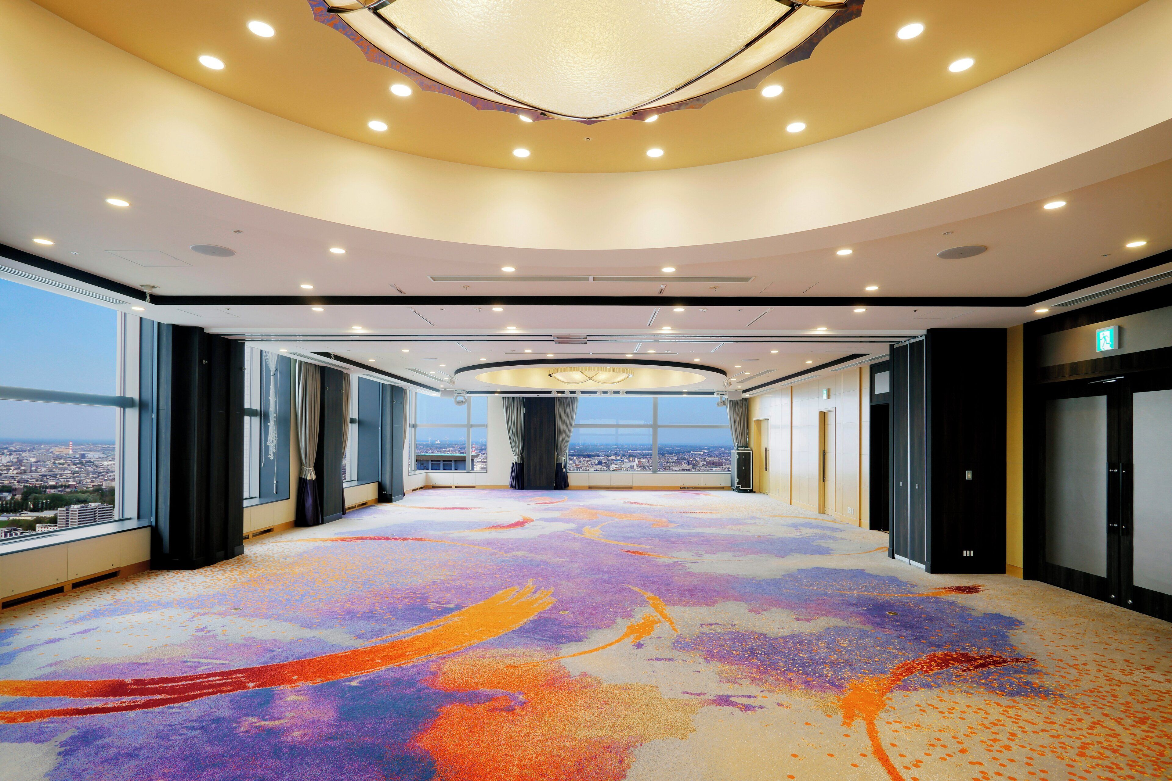 A spacious hotel lobby with a colorful carpet, large windows offering city views, and a modern ceiling design.