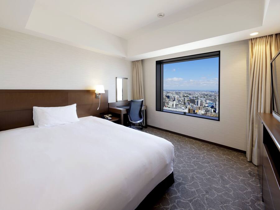 Habitación de hotel con una cama grande, un escritorio con silla, televisor y ventana con vista a la ciudad.