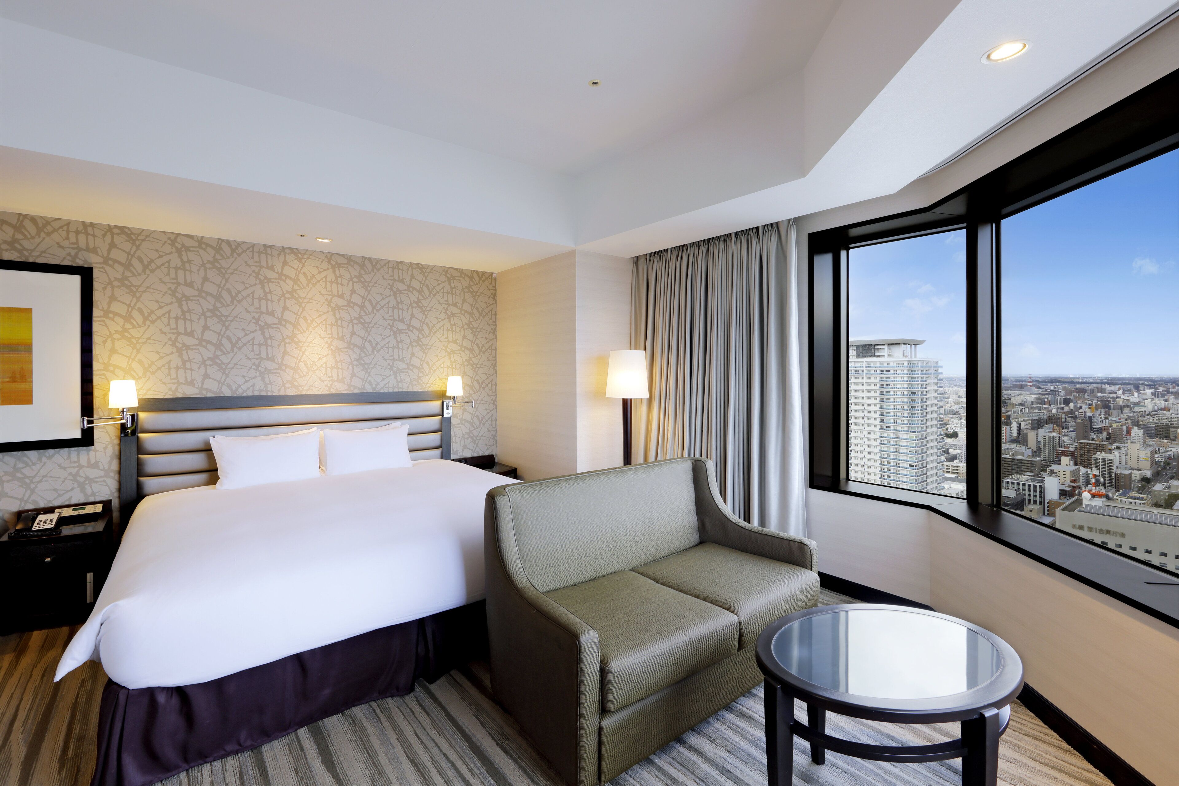 Habitación de hotel con una cama grande, un sofá, una mesita y vistas a la ciudad a través de amplios ventanales.