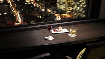 A cityscape view from a window with a table featuring a glass of red wine, a glass of whiskey, and a dessert plate.