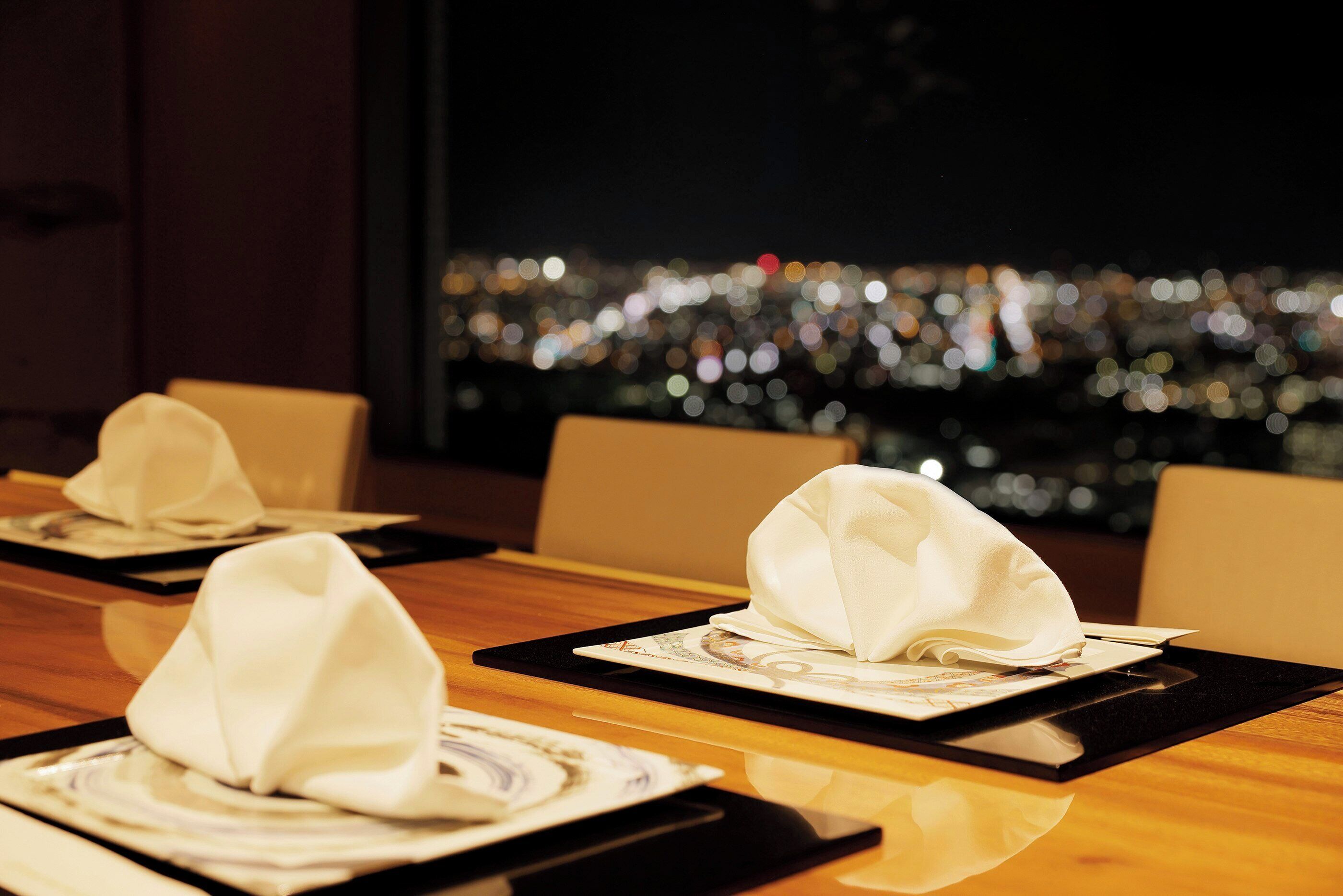 Mesa de jantar com pratos e guardanapos arrumados, refletidos na superfície da mesa, com vista para a cidade ao fundo.