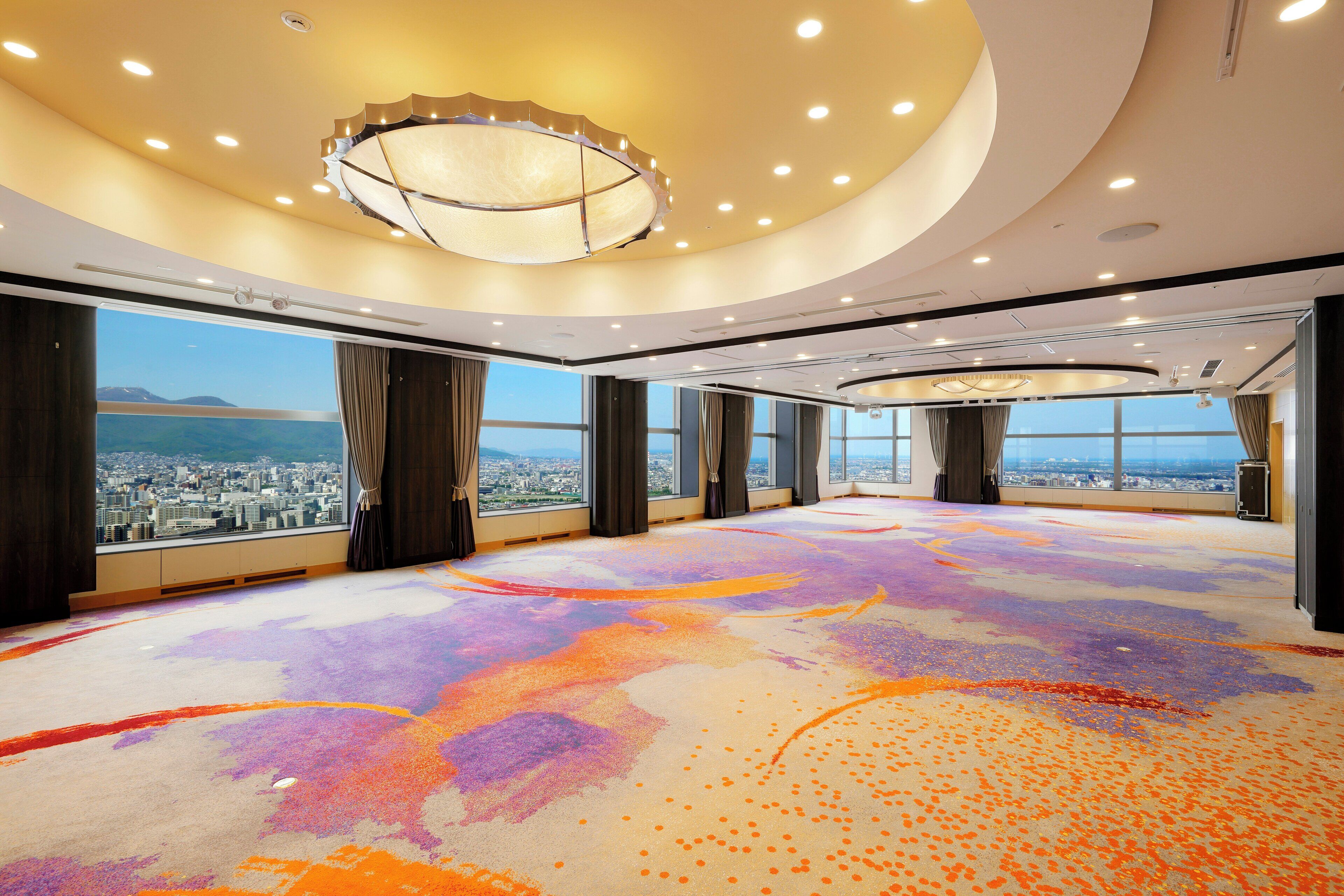 A spacious room with a panoramic city view, a colorful carpet, and a modern chandelier.