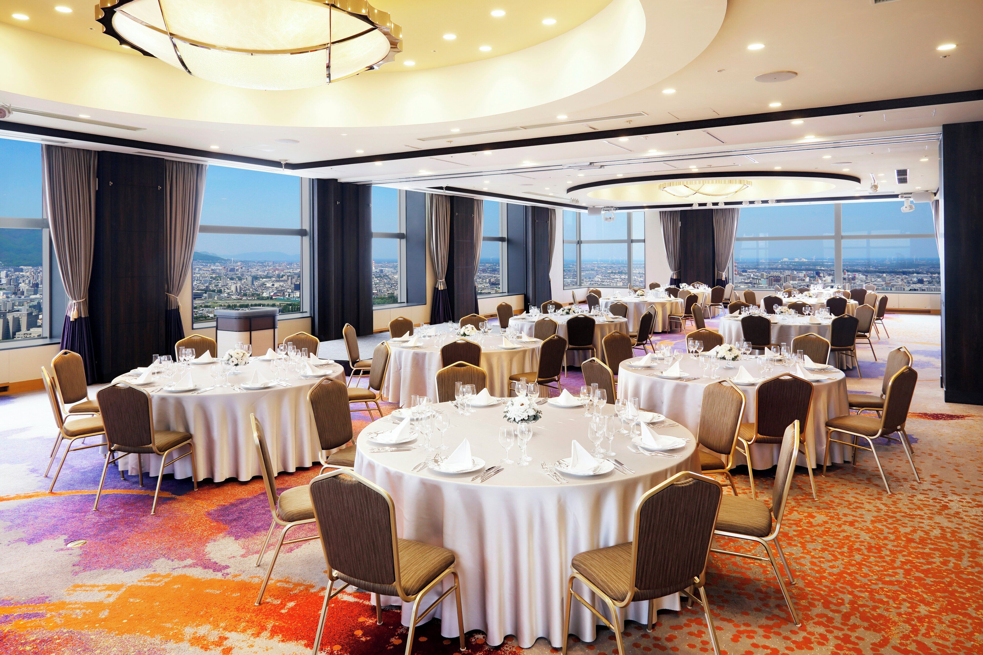 A conference room with multiple round tables, each set with white tablecloths, chairs, and glassware. The room features large windows with a cityscape view.