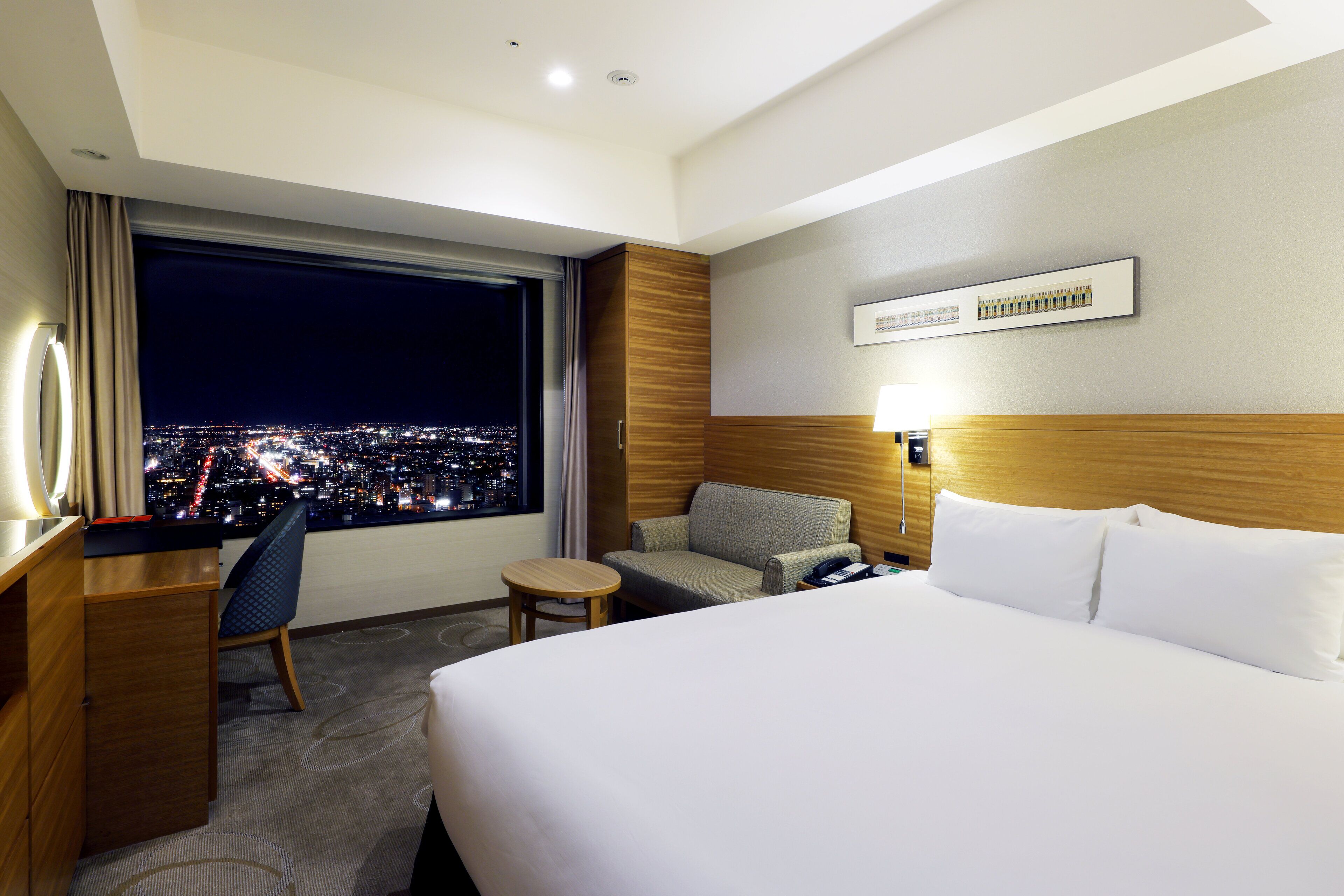 A hotel room with a large bed, a desk, a chair, and a view of a cityscape at night.