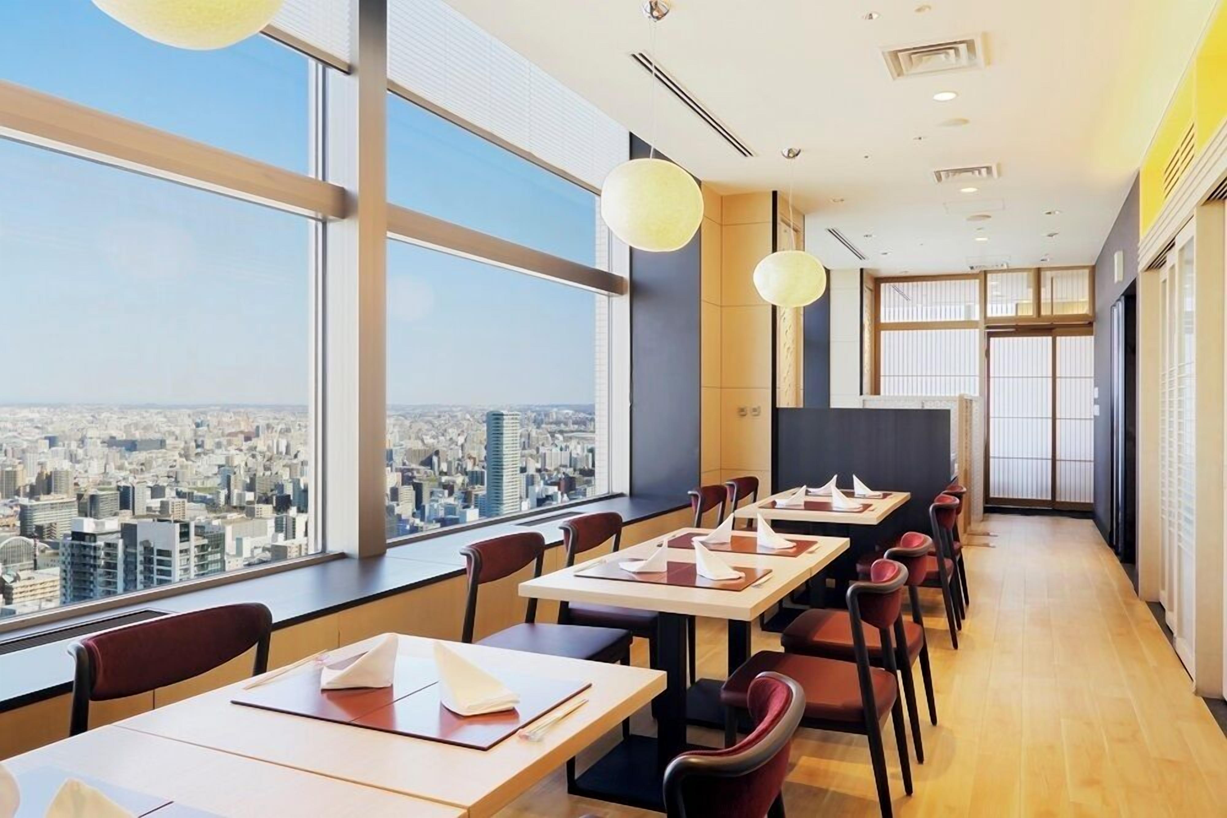 Un comedor moderno con mesas y sillas, ventanas amplias con vista a la ciudad e iluminación colgante.