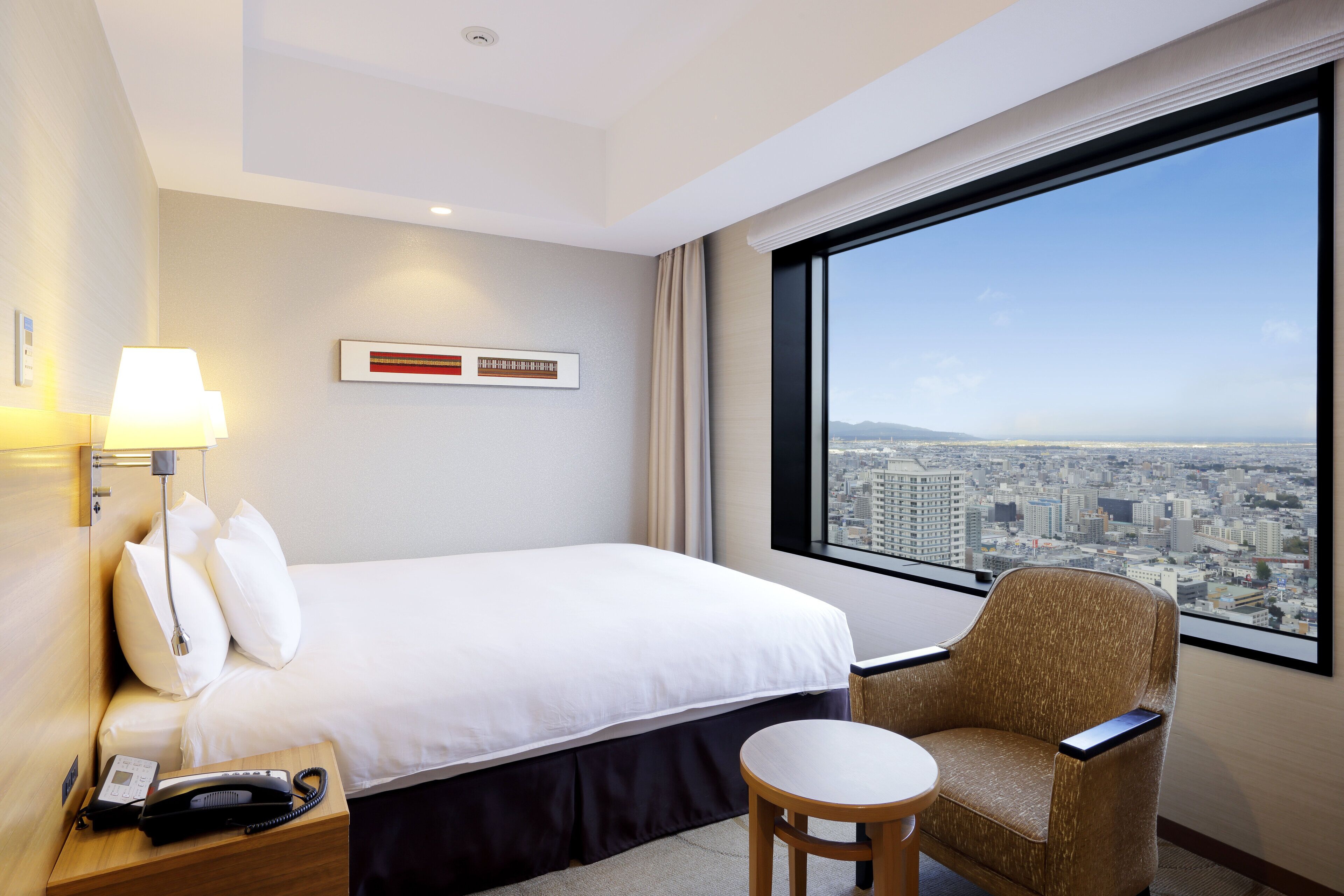 A hotel room with a bed, a chair, a small table, a lamp, and a phone on a nightstand. There is a large window with a city view.