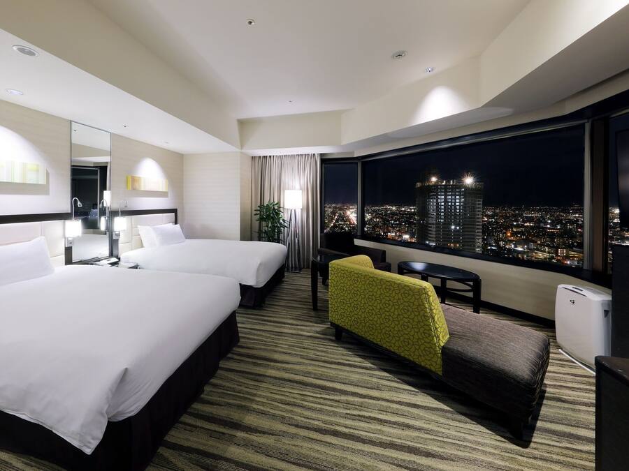 Habitación de hotel con dos camas, un sofá, una mesita y un amplio ventanal con vistas a la ciudad.