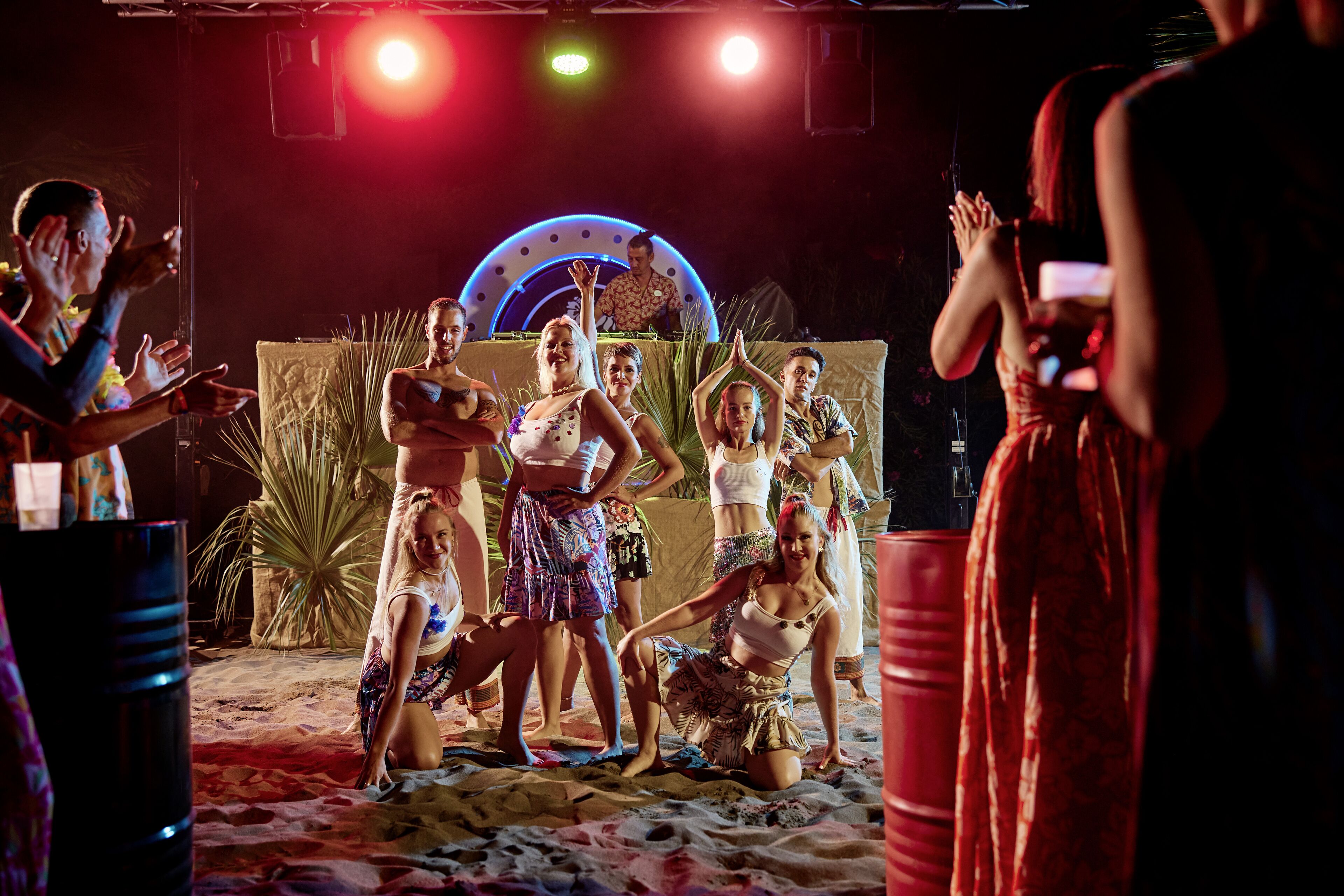 A group of people dancing on a sandy stage with a large circular light structure in the background.