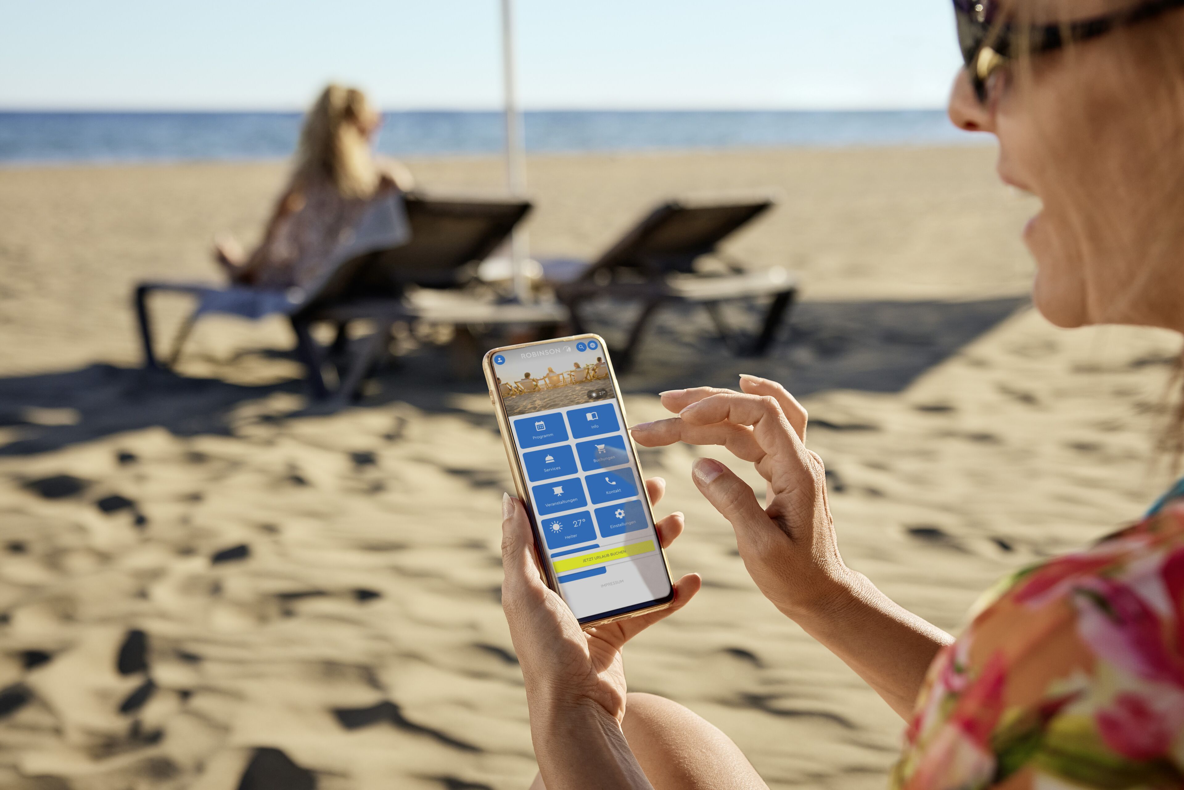 A person using a smartphone on a beach with lounge chairs and a clear view of the ocean.
