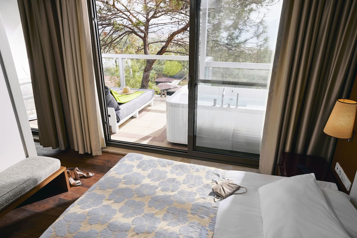 Ein Hotelzimmer mit Bett, einer Bank, einem kleinen Tisch und einer sichtbaren Whirlwanne durch die Glastür.